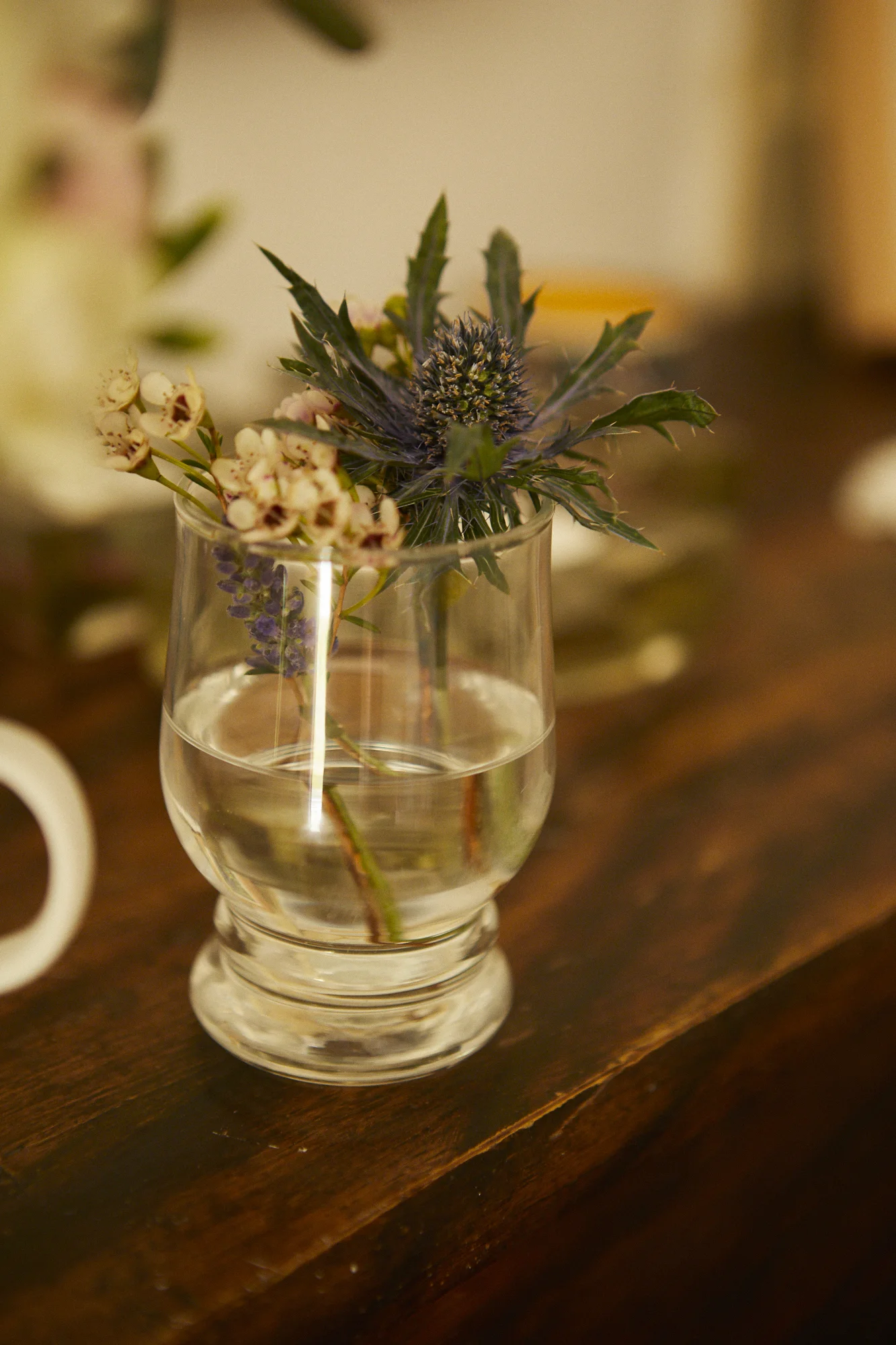 A glass vase with water containing assorted flowers and foliage, placed on a wooden surface.