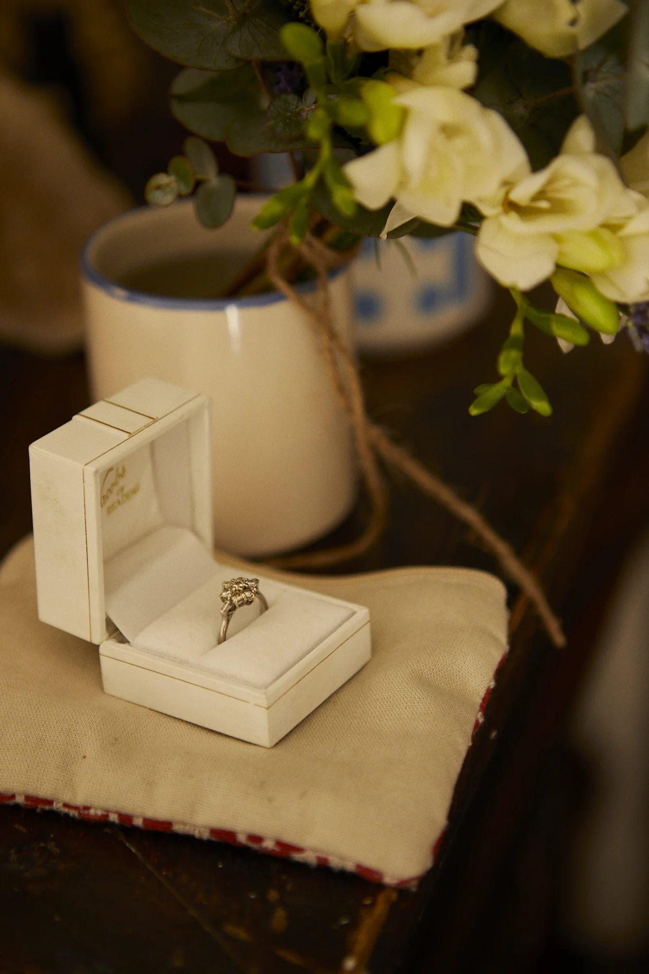 An engagement ring in a white jewelry box, placed on a beige cloth, with a white vase of white flowers in the background.
