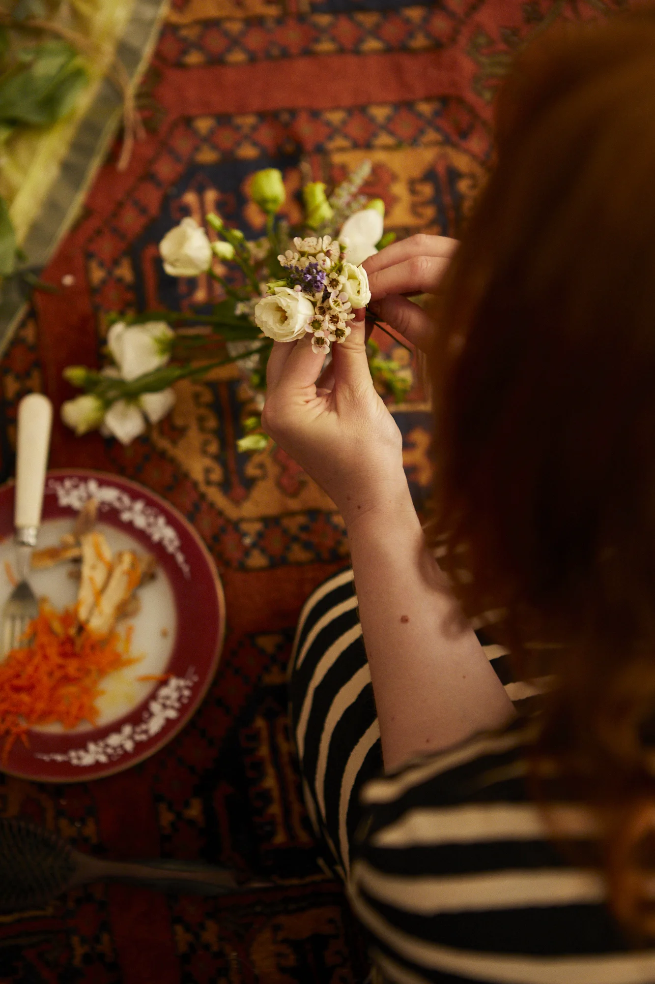 A person sitting on a patterned rug, holding a small bouquet of white and purple flowers, with a plate of partially eaten food and a fork on the side.