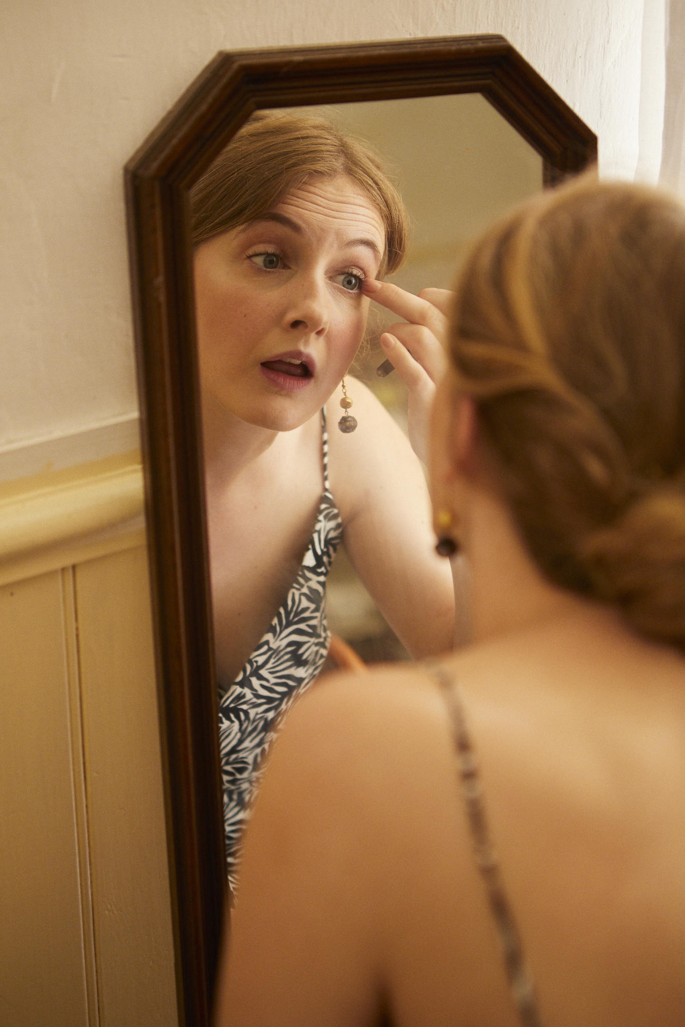 A woman with red hair applying makeup in front of a mirror.
