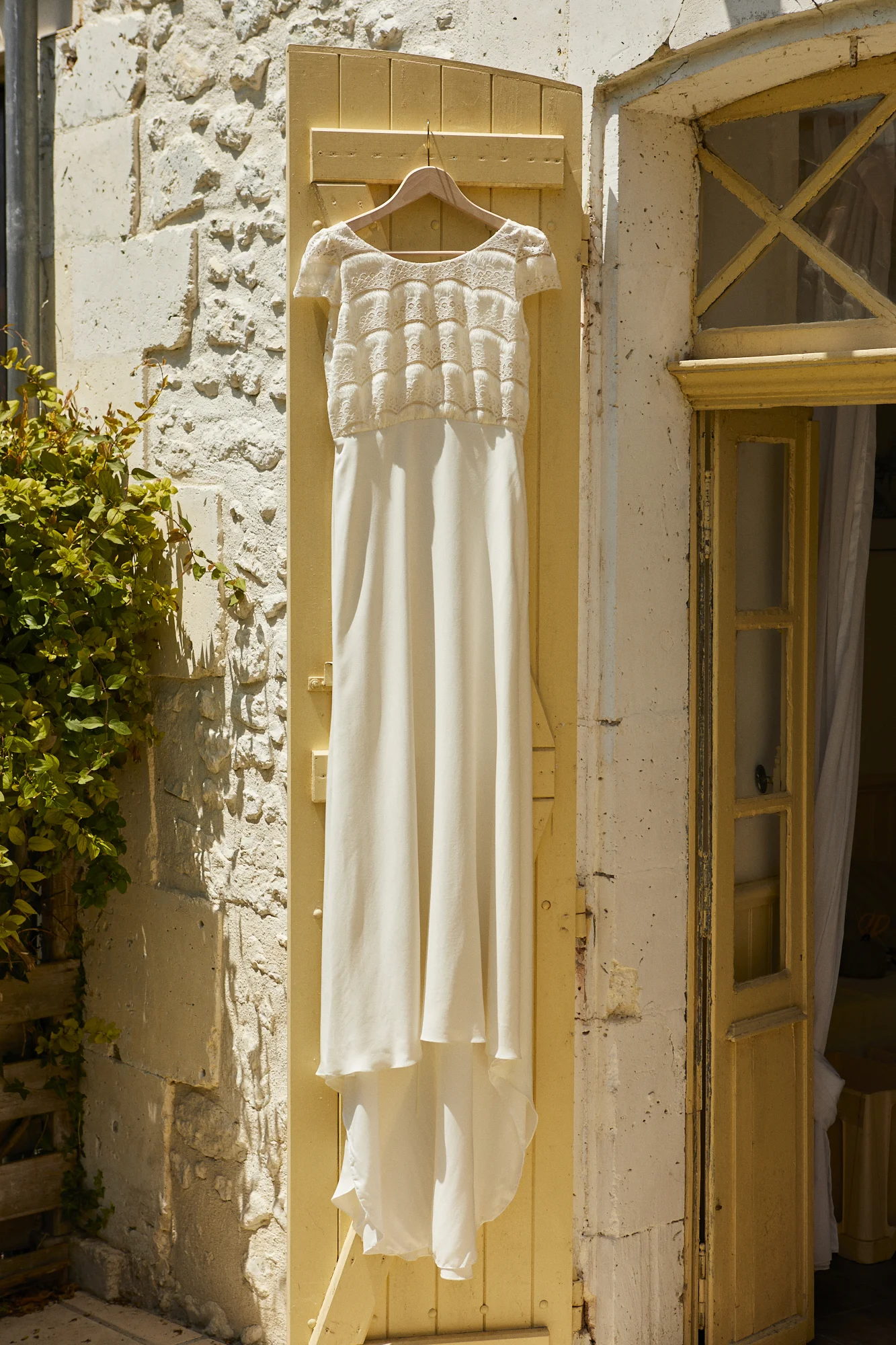 A white dress hanging on a hanger outside on a yellow door, with a stone wall and an open window with yellow trim in the background.