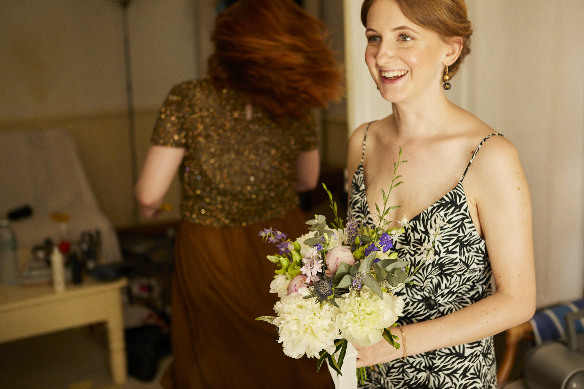A woman with short red hair wearing a black and white patterned slip dress holding a bouquet of flowers and smiling indoors. Another woman with red hair is in the background, facing away.
