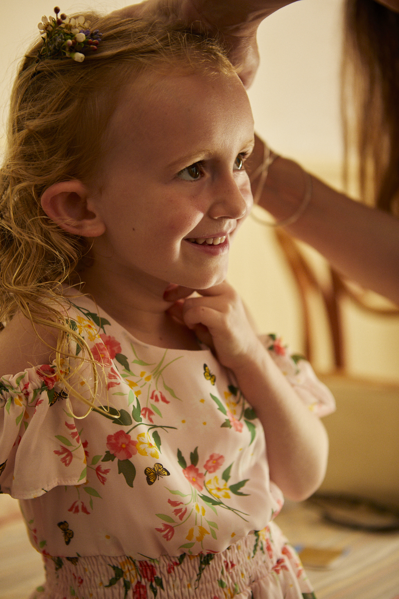 A young girl with curly blonde hair, wearing a floral dress, smiling as someone adjusts her hair accessories.