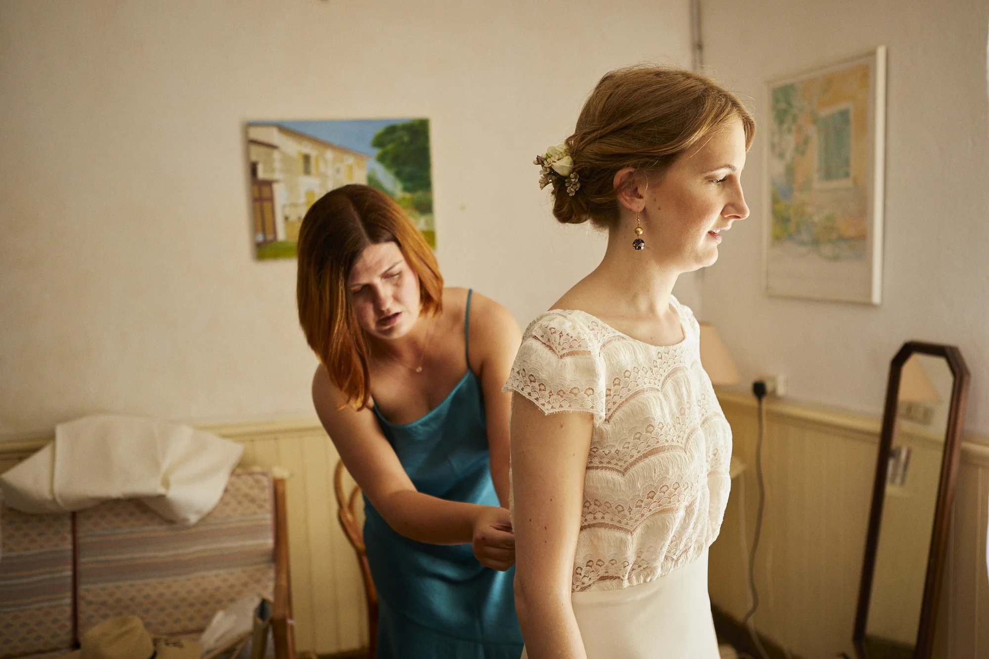 A bride getting ready, with a woman helping her with her dress, in a room decorated with paintings on the wall.