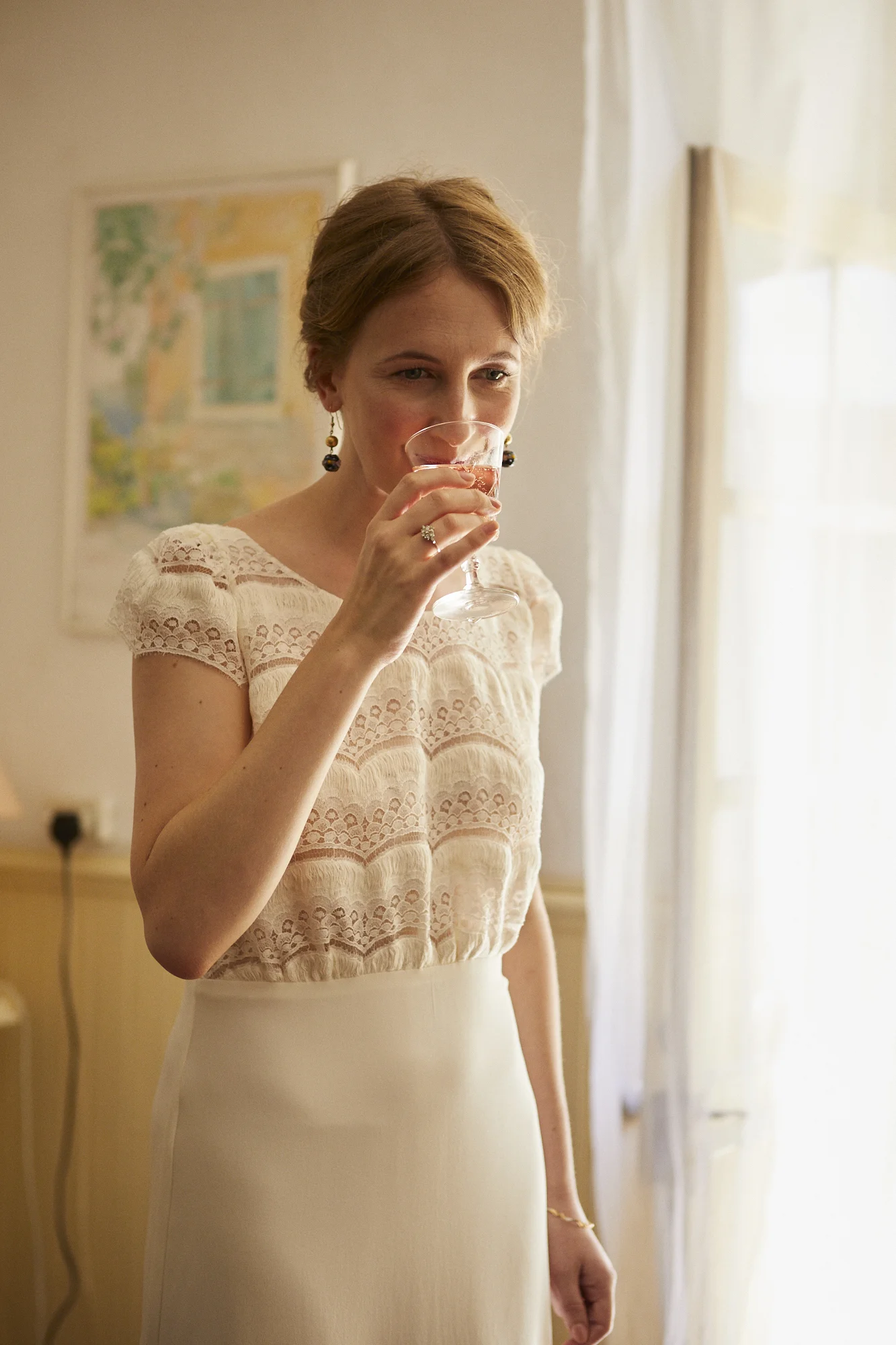 A woman with short, light brown hair in a vintage-style, cream-colored dress with lace on the top, holding a glass of pink wine and smiling softly.