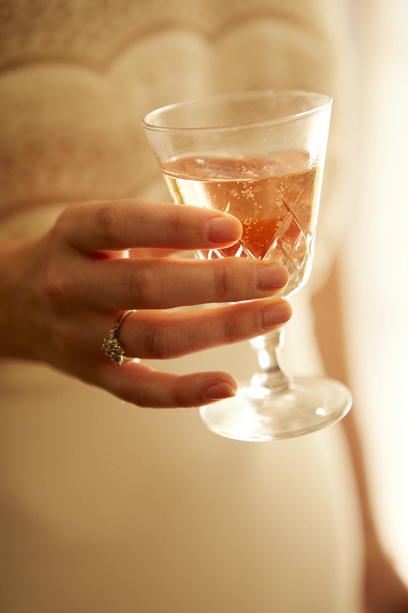 A hand holding a glass of rosé wine or champagne with a diamond ring on the ring finger, against a warm, blurred background.