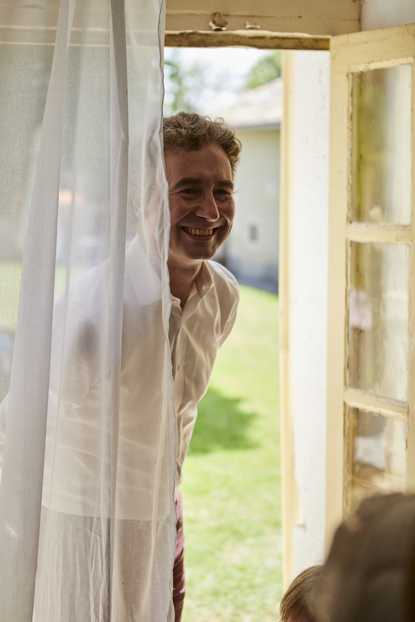 A man with curly hair and a big smile, wearing a white shirt, peeking through a sheer white curtain from outside a house.