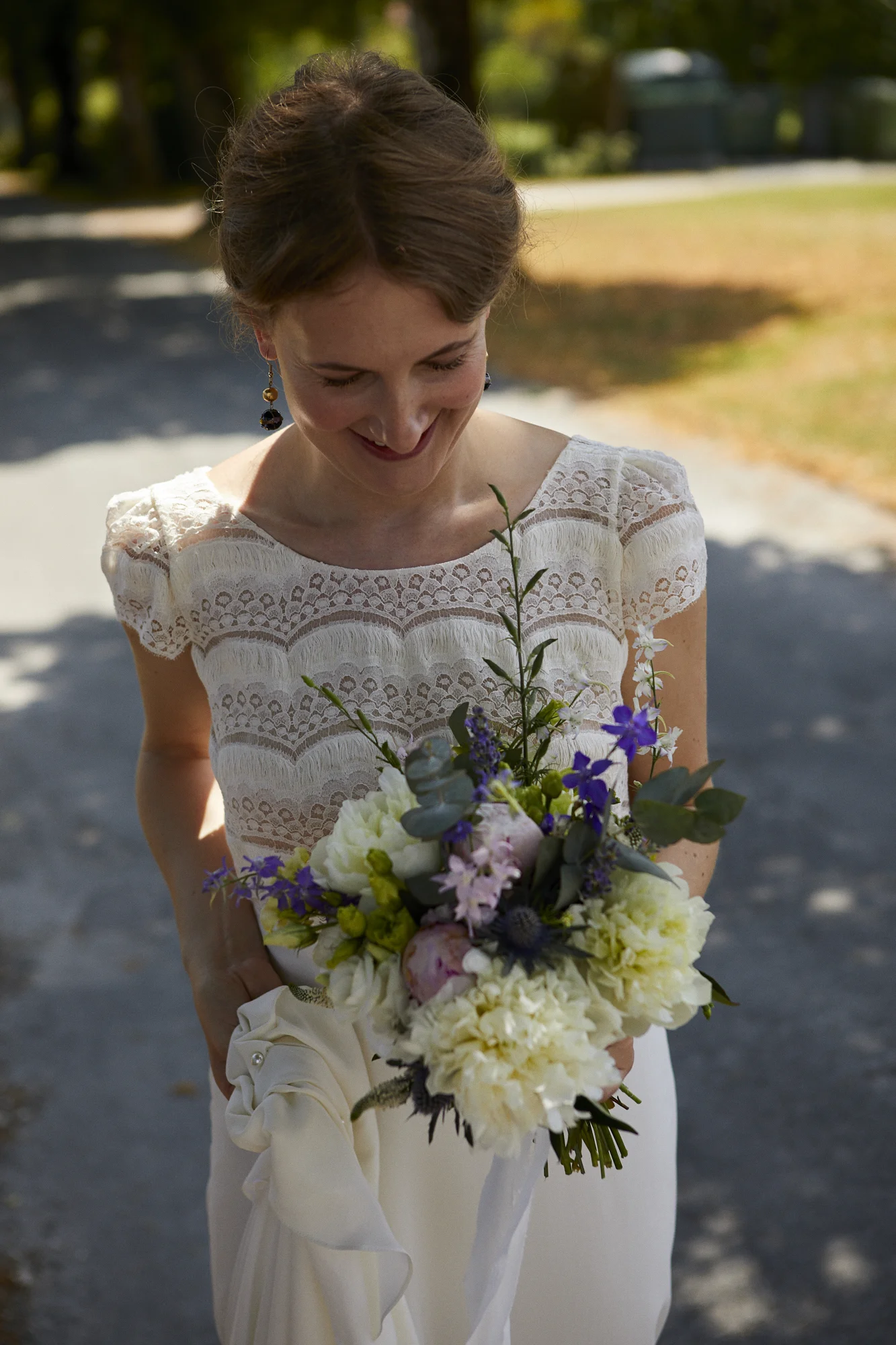 A woman in a white lace dress holding a bouquet of flowers, standing outdoors on a sunny day.