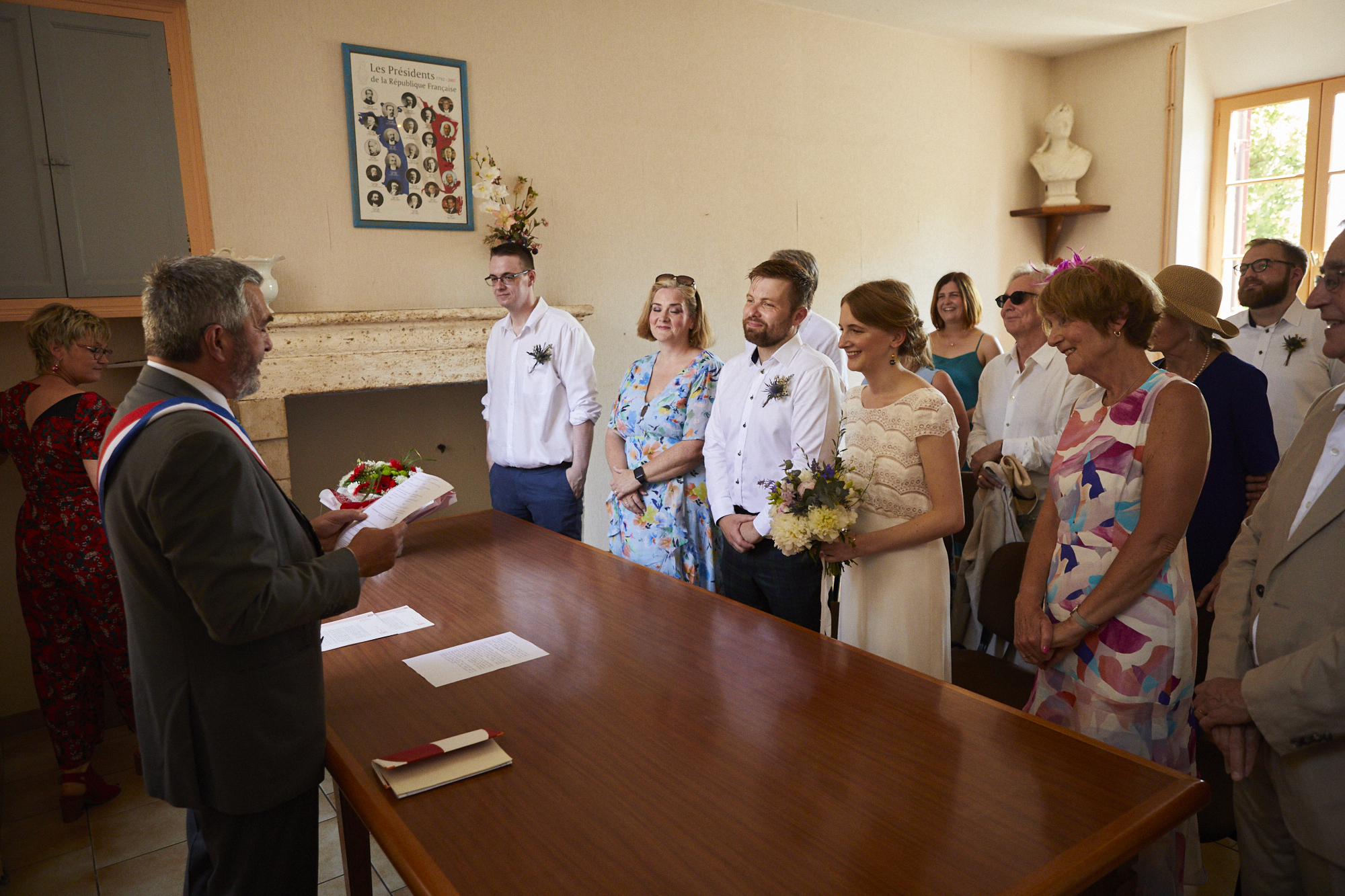 A wedding ceremony taking place indoors. The officiant reading from a paper stands at a table, with the bride holding flowers and couples and family members watching and smiling.