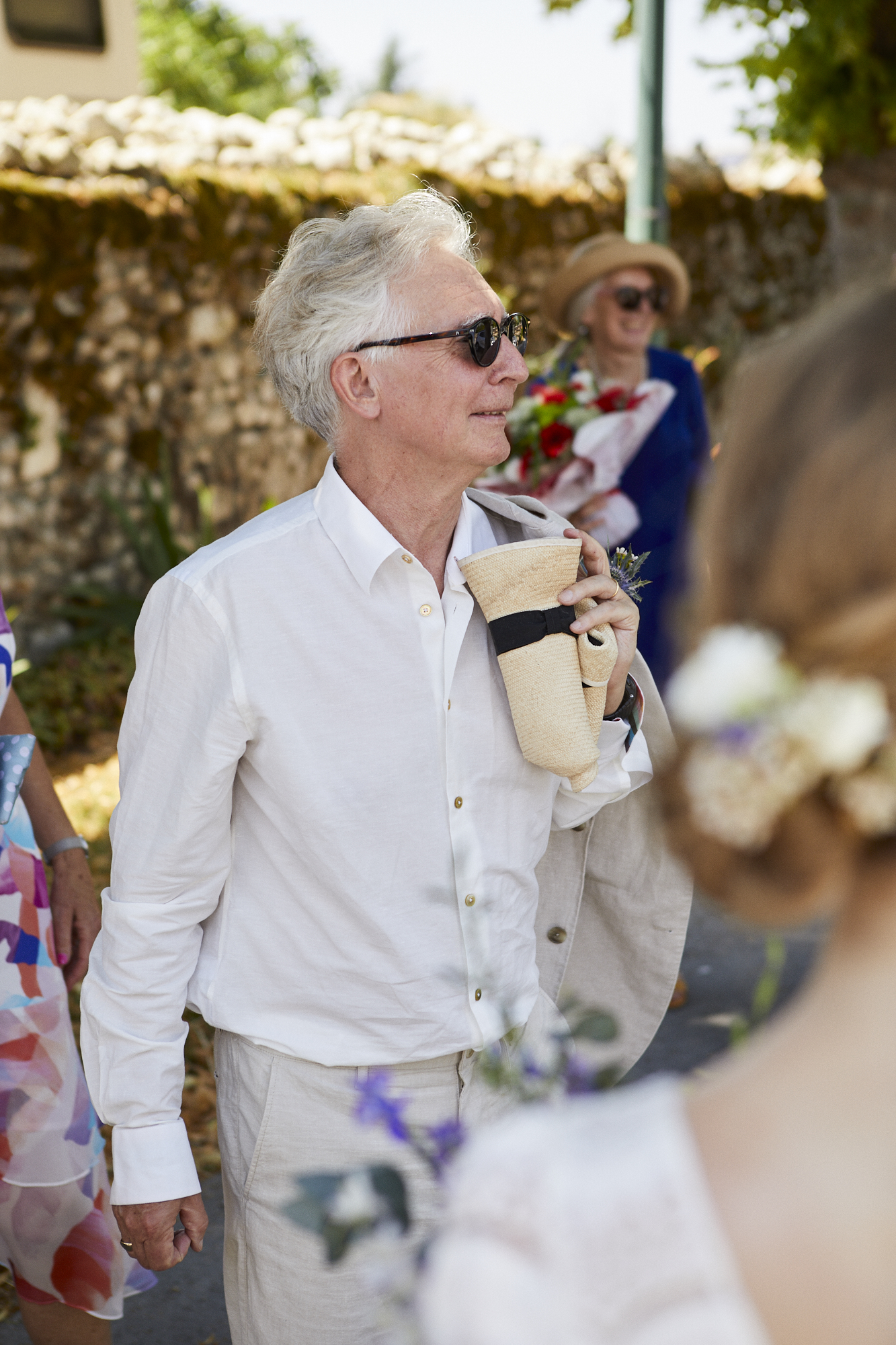 An elderly man with gray hair, wearing sunglasses and a white shirt, holding a beige hat and a jacket over his shoulder, stands outdoors during the daytime with a woman in the background holding a bouquet of flowers.