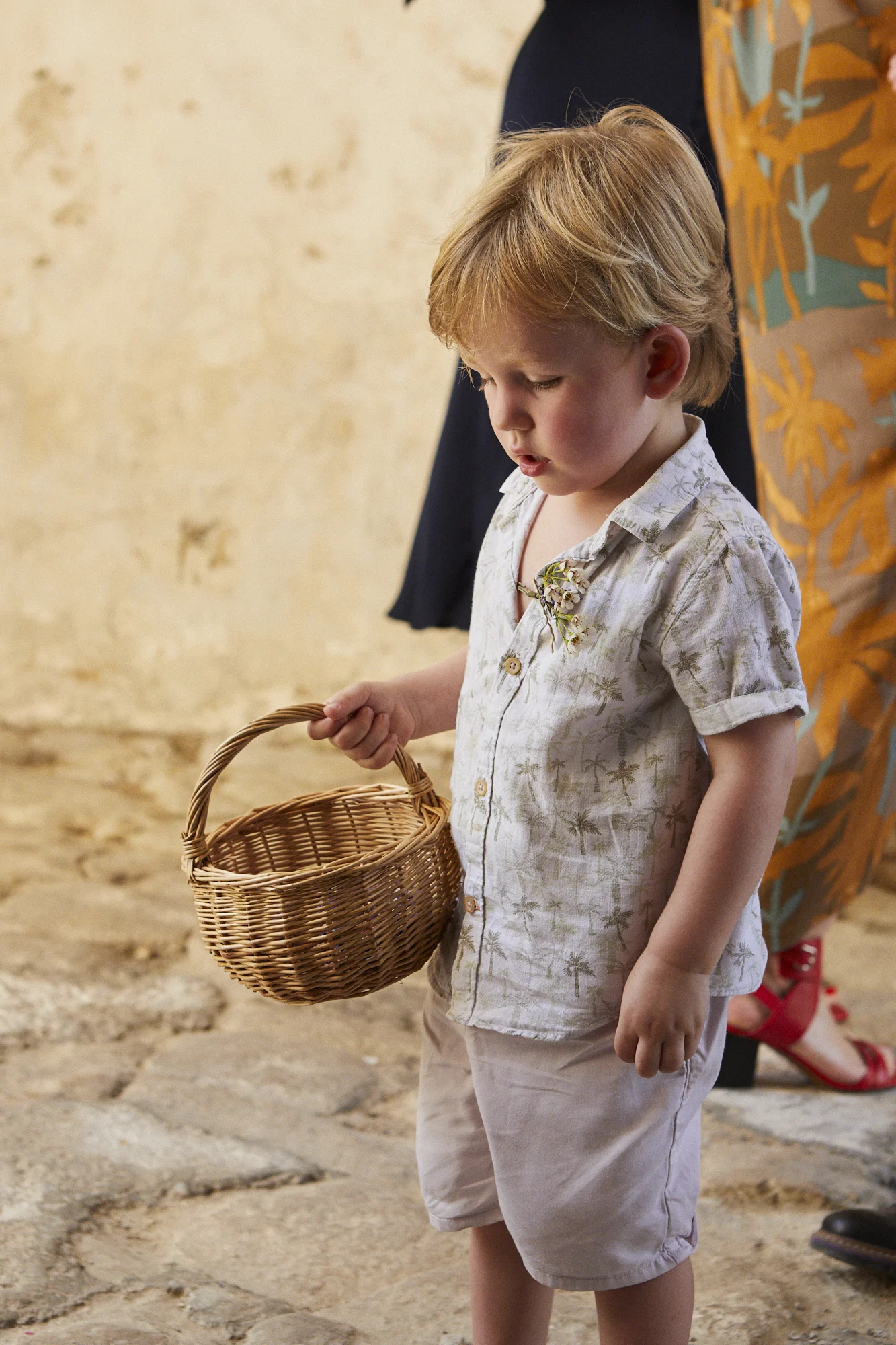 A young boy with blond hair holding a small wicker basket, looking down thoughtfully, standing on a stone floor, with two women partially visible behind him.
