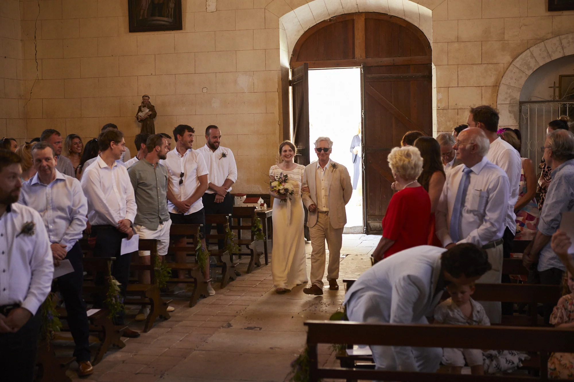 A bride and her father walking down the aisle in a church decorated for a wedding, with guests standing and watching.