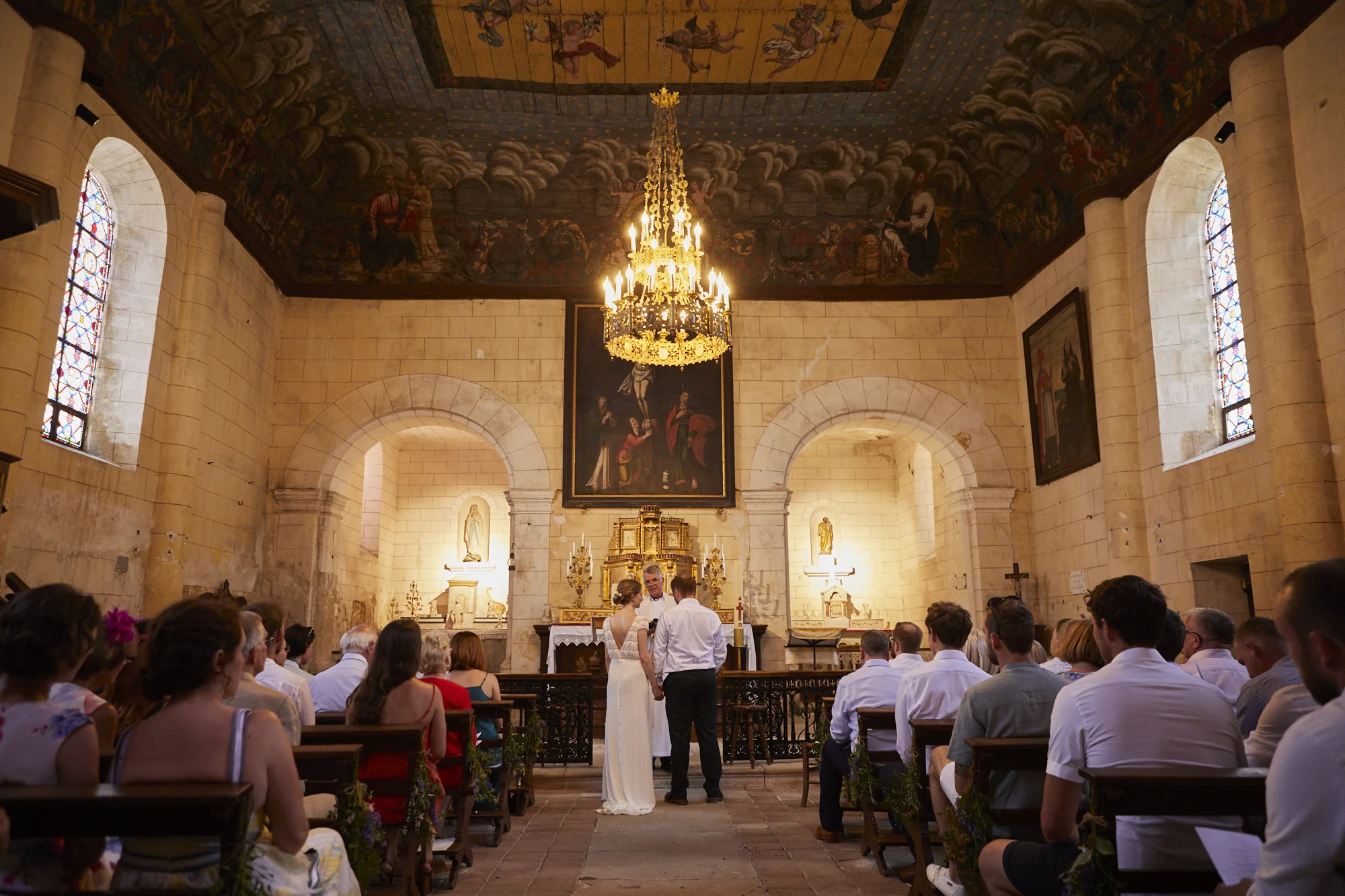 A wedding ceremony inside a historic church with stone walls, stained glass windows, and religious artwork. The bride and groom stand before the officiant, with guests seated on wooden benches facing them. A chandelier hangs from the ceiling, and sta