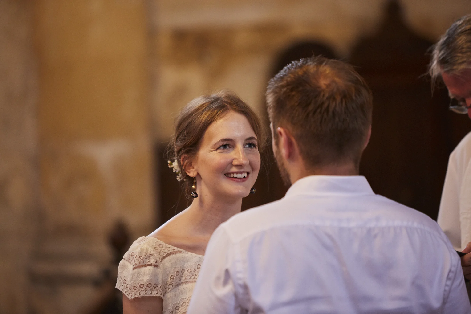 A woman with brown hair and earrings, smiling at a man with short brown hair, during a wedding or similar ceremony.