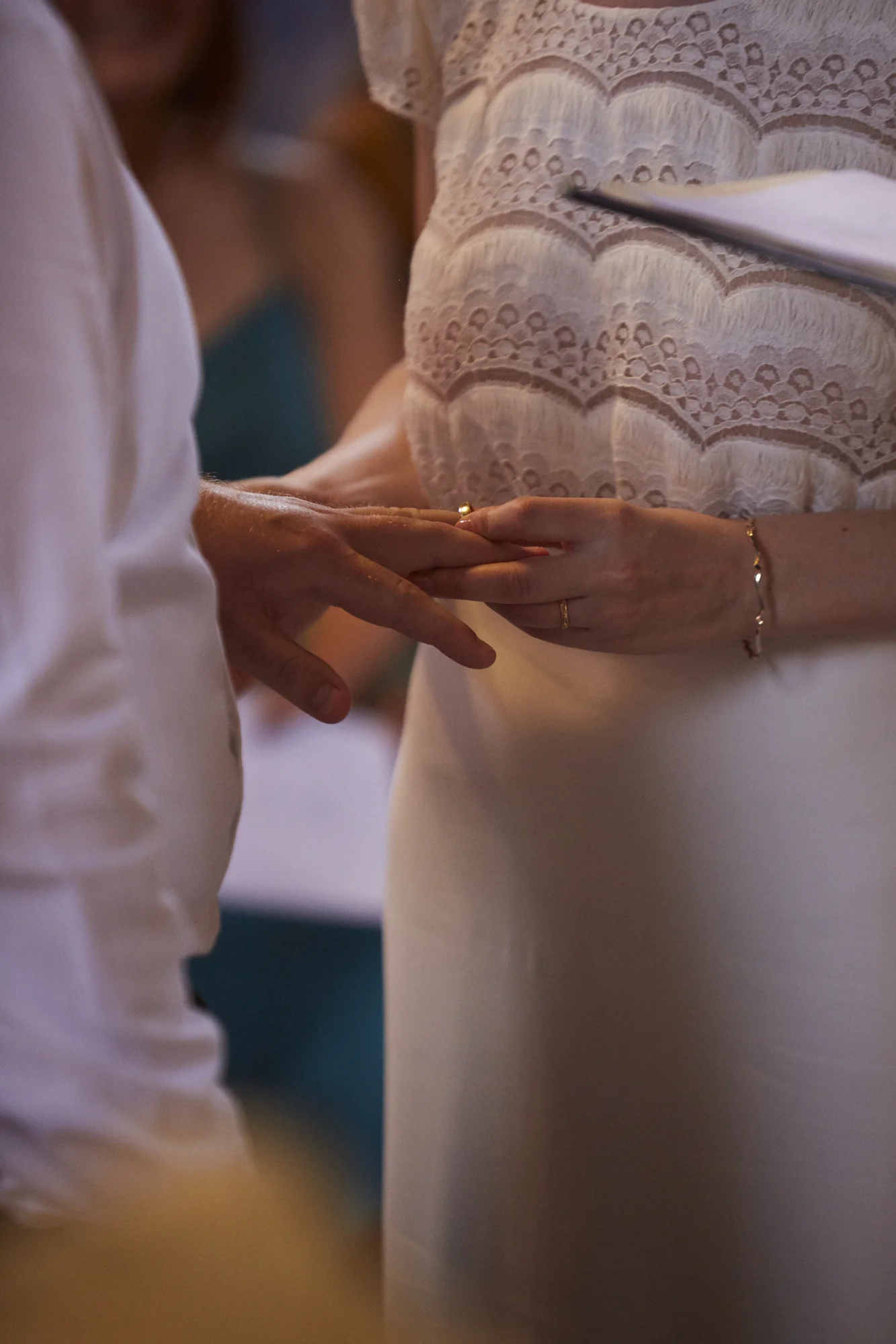 Close-up of a couple holding hands during a wedding ceremony, with the bride's hand resting on the groom's hand, both wearing wedding rings, and the bride wearing a lace dress and jewelry.
