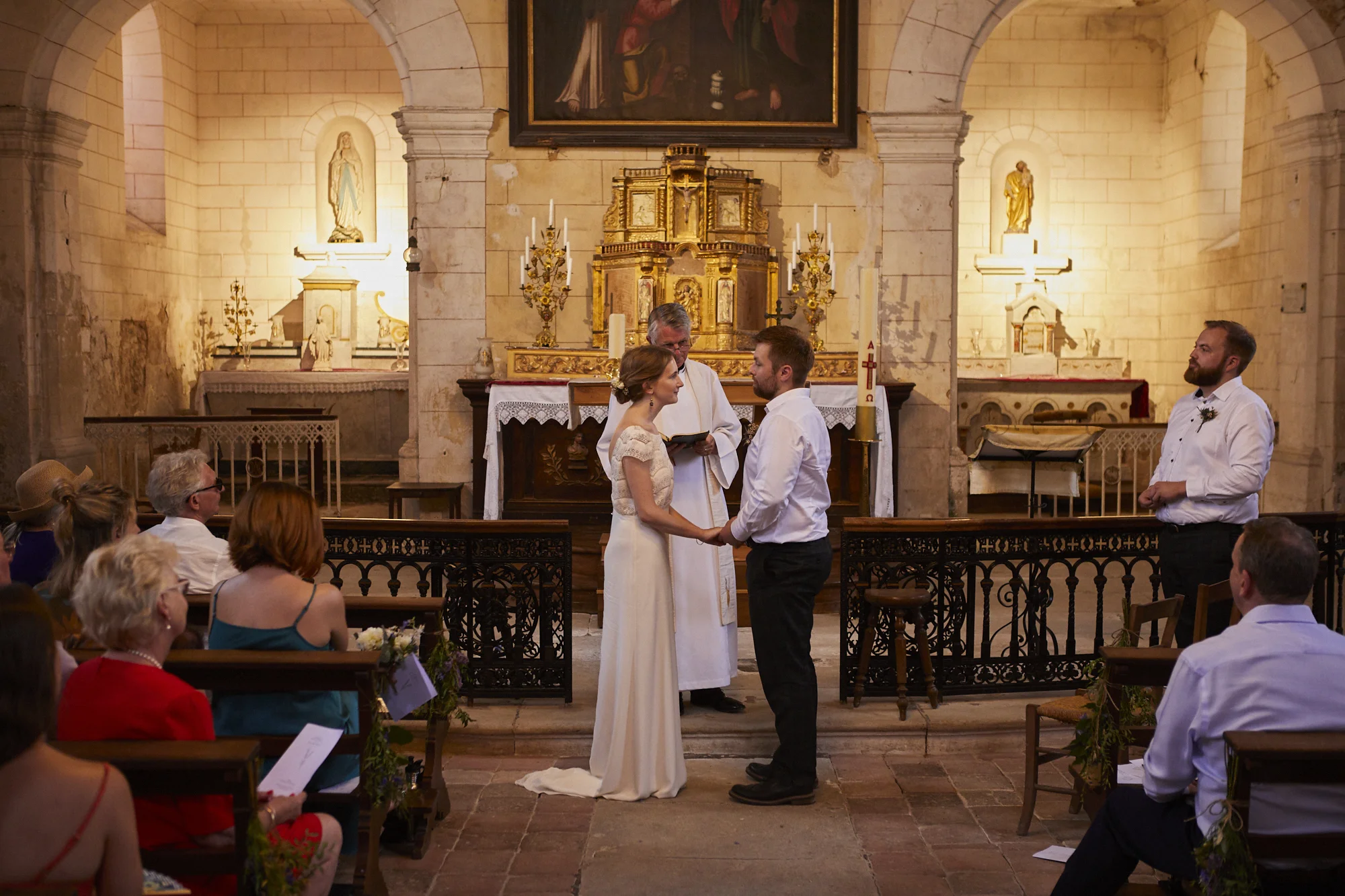 A wedding ceremony taking place inside a church. The bride and groom are holding hands and facing each other, with a priest standing behind them, reading from a book. Guests are seated, watching the ceremony. The church has an ornate altar with gold 
