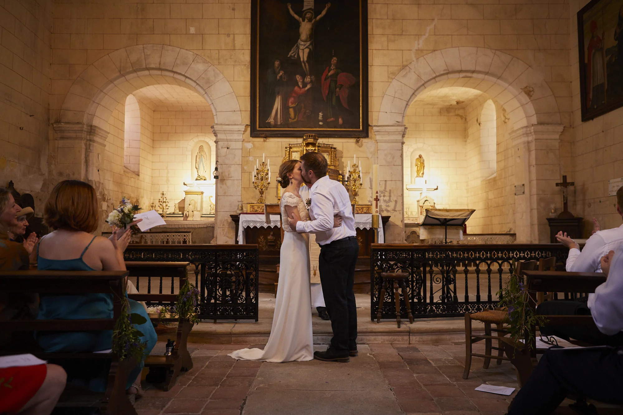 A bride and groom kissing at their wedding ceremony inside a church, with guests clapping and taking photos, and religious artwork on the walls behind them.