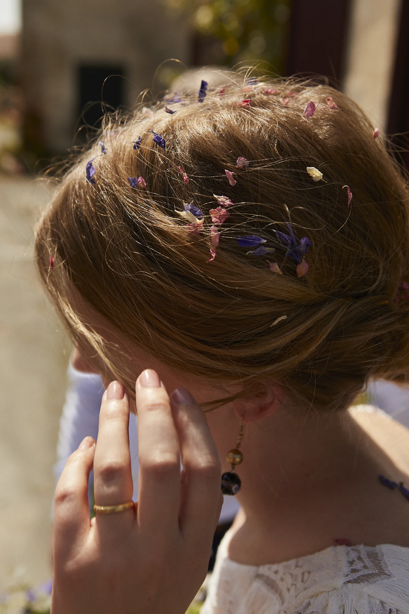 Close-up of a woman with red hair decorated with purple, pink, and white flower petals, touching her face with her hand, outdoors with sunlight and blurred background.