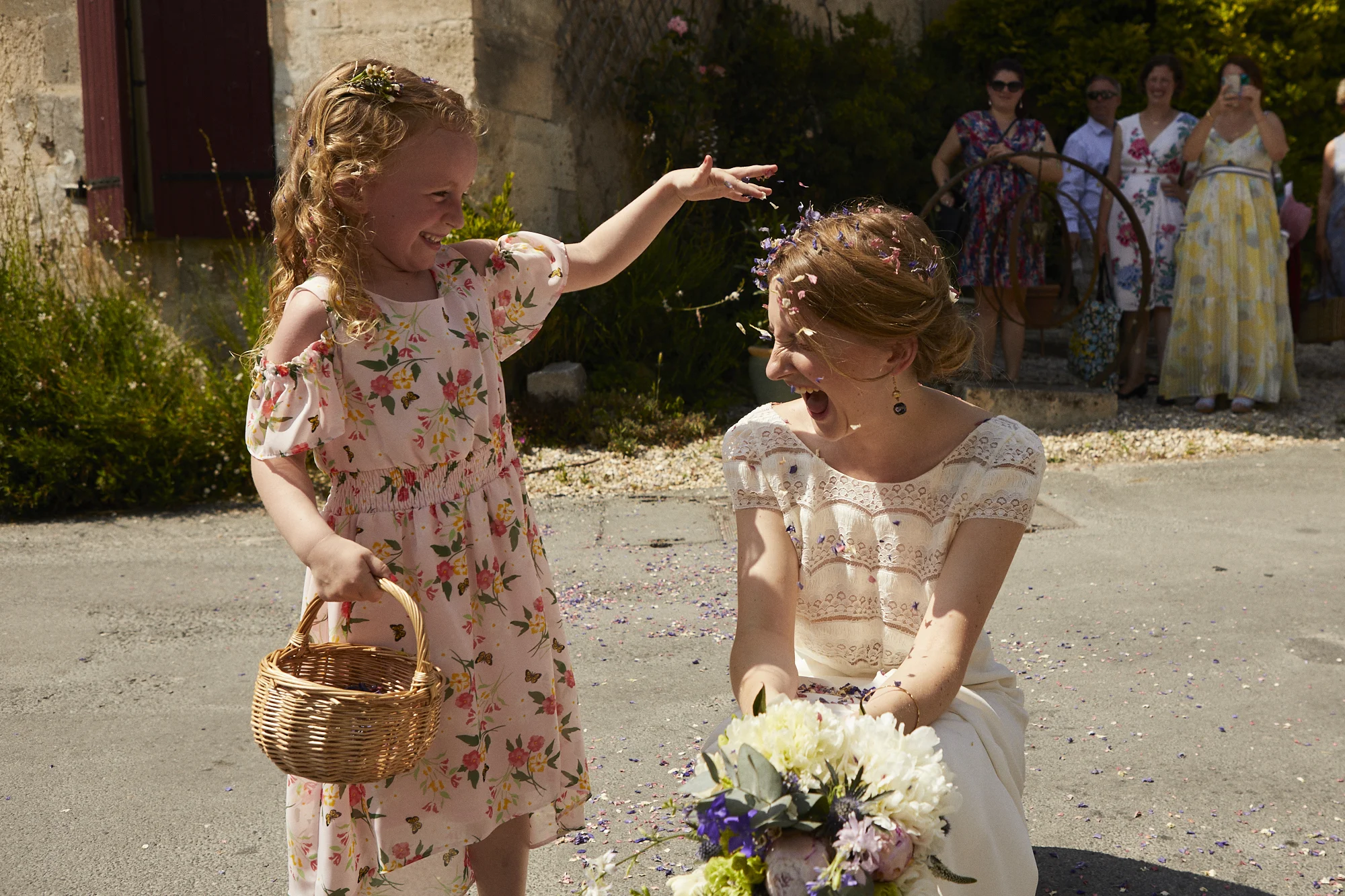 A woman in a white dress is kneeling with a bouquet of flowers, reacting happily as a young girl in a pink floral dress sprinkling confetti on her during an outdoor celebration, with other women in dresses watching.