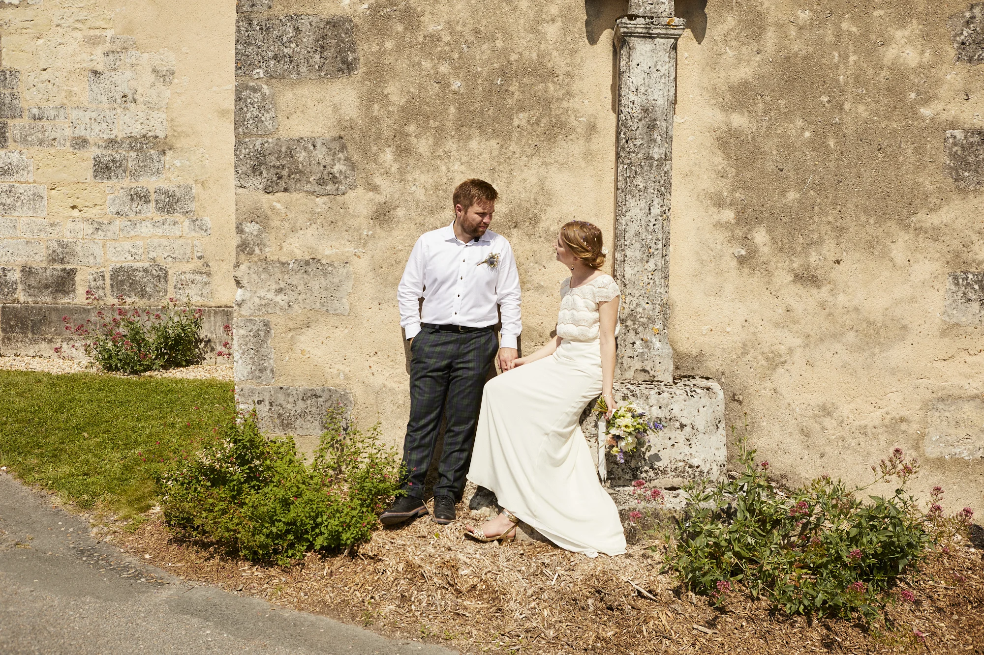 A couple in wedding attire standing near a stone wall, with the woman sitting on a stone ledge holding a bouquet, as they look at each other.
