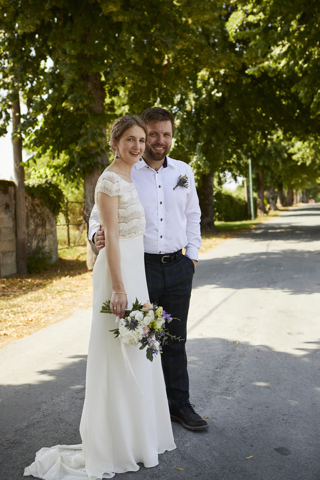 A newlywed couple standing on a tree-lined street, smiling at the camera. The bride is holding a bouquet of flowers, and the groom has his arm around her waist.
