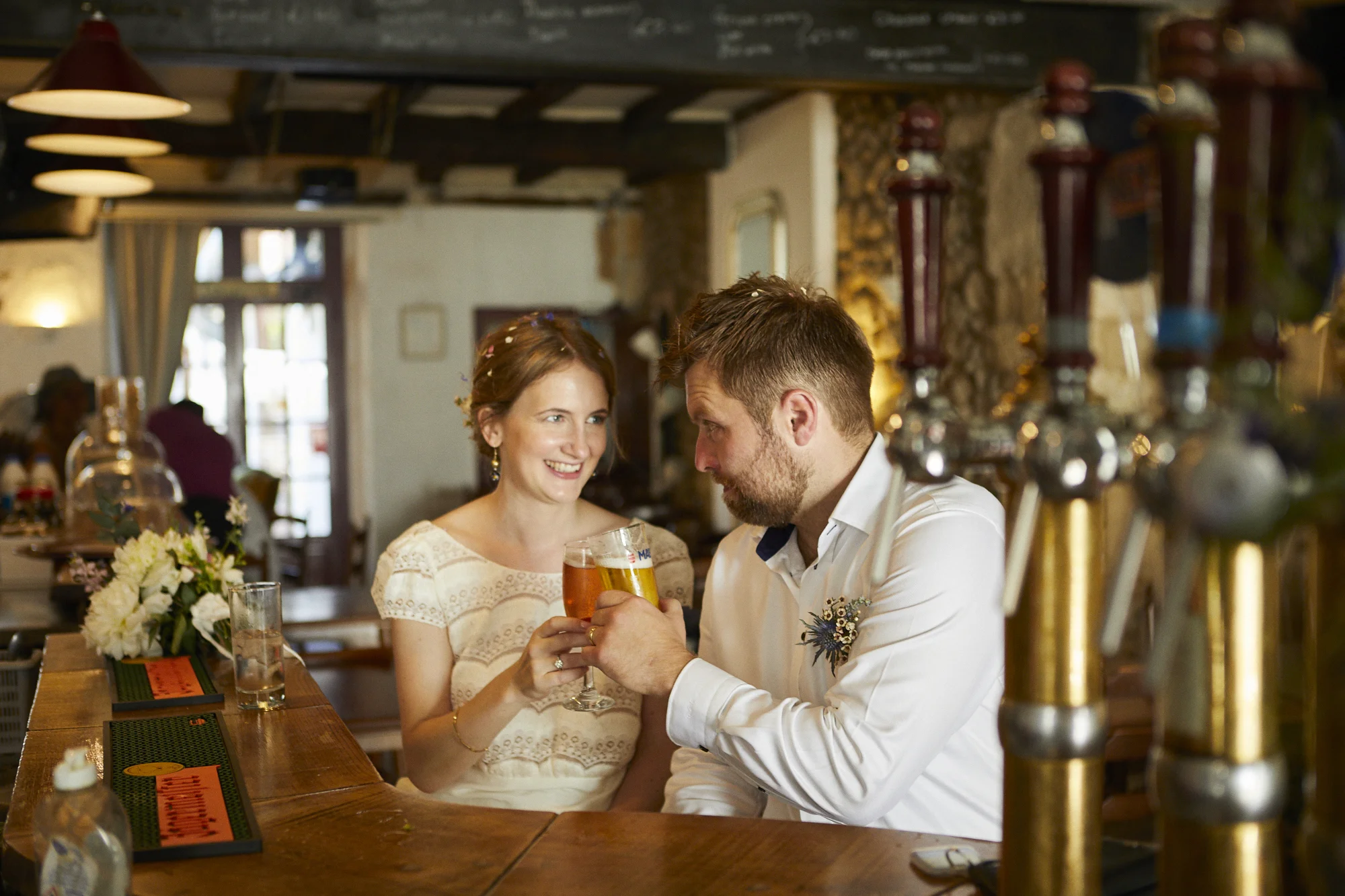A smiling woman and a man clinking glasses of beer at a bar in a cozy restaurant or pub, with floral arrangements and decorative pipes in the foreground.
