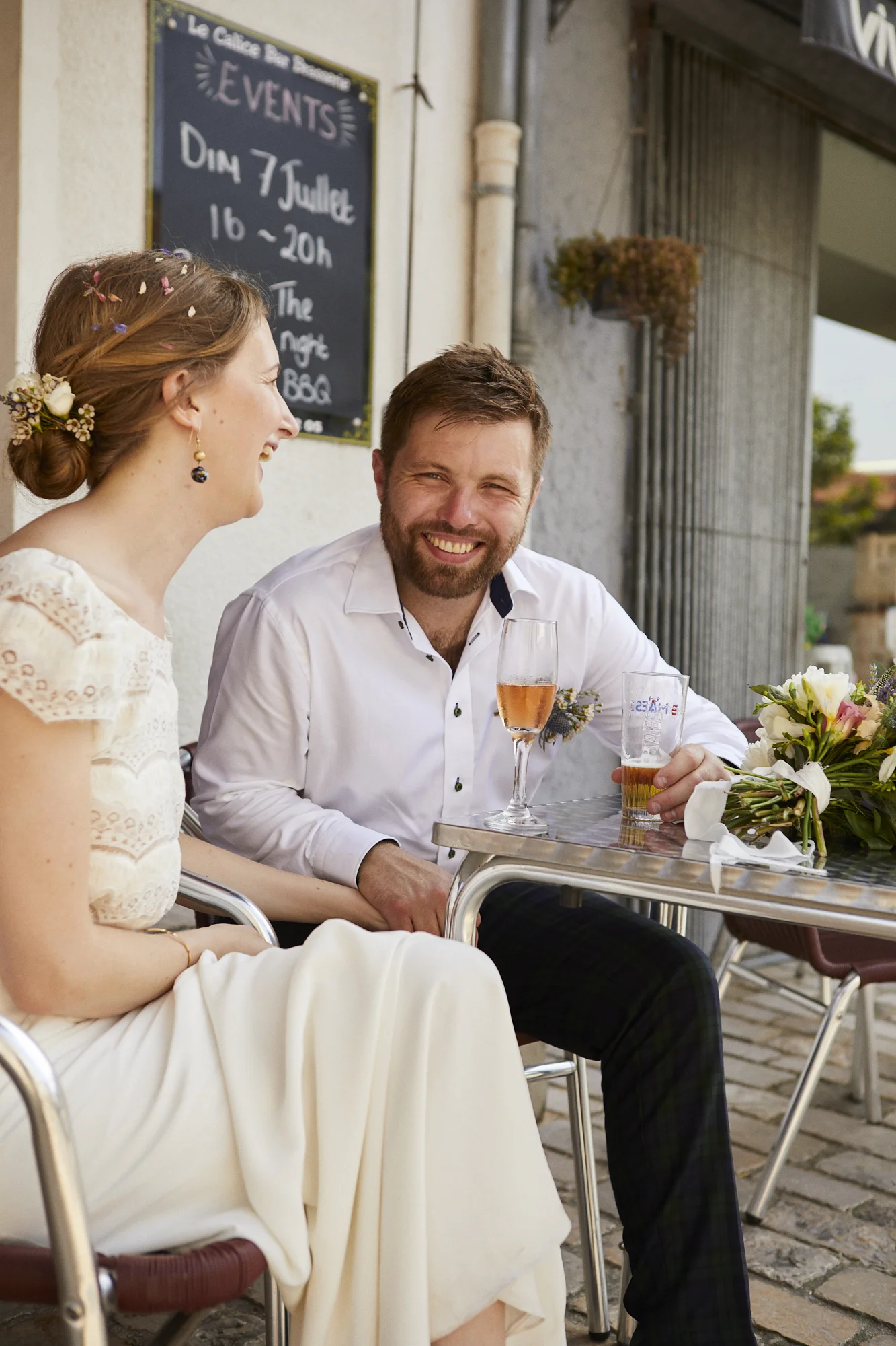A man and woman are seated at an outdoor table, smiling and enjoying drinks, with a bouquet of flowers on the table. The woman wears a white dress with lace details and has her hair styled with flowers and tiny ribbons. The man has a beard, wears a w