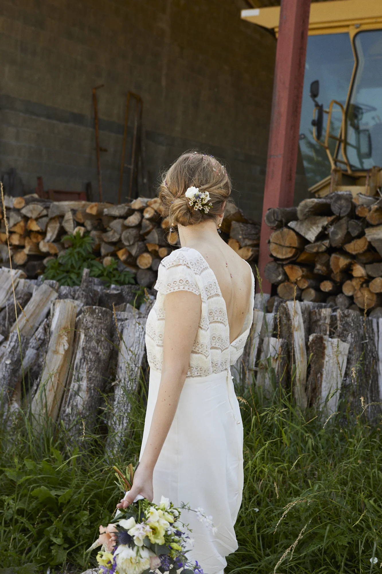 A woman in a white dress holding a bouquet of flowers, standing outdoors with firewood stacked behind her.