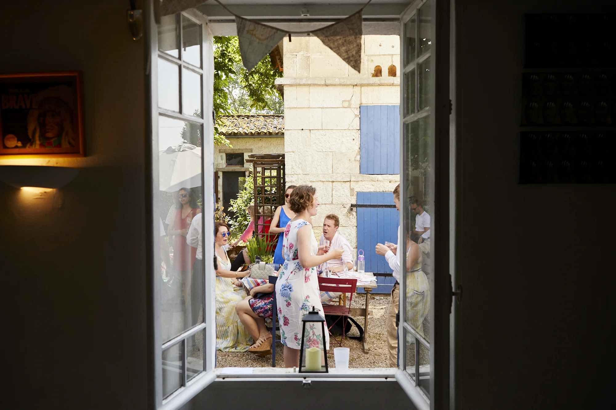 View through a window looking out onto a garden gathering with people chatting, standing, and sitting at tables during daytime.