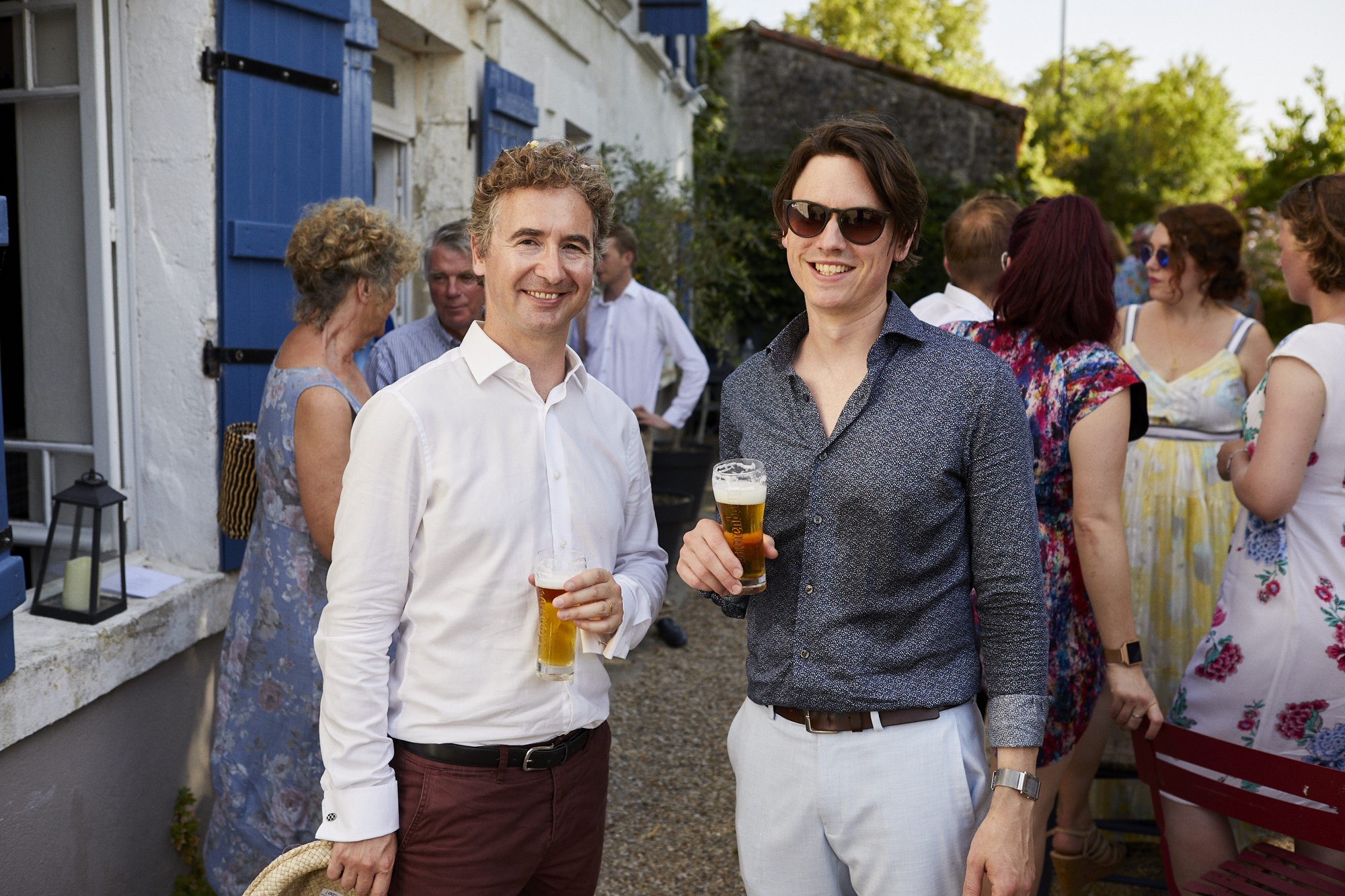 Two men smiling and holding glasses of beer at an outdoor social gathering. There are other people in the background, talking and enjoying the event, with a building featuring blue shutters behind them.