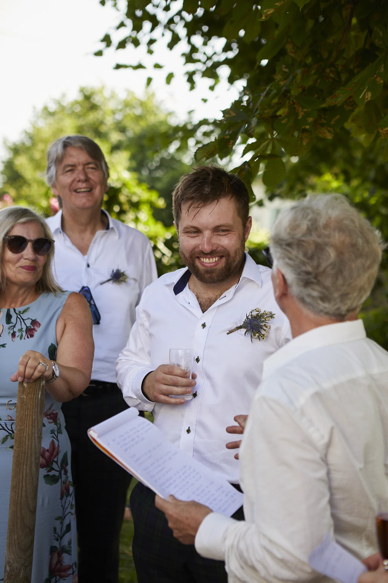 People at an outdoor gathering, some wearing sunglasses and holding drinks, with a man reading from a paper, under a tree canopy.