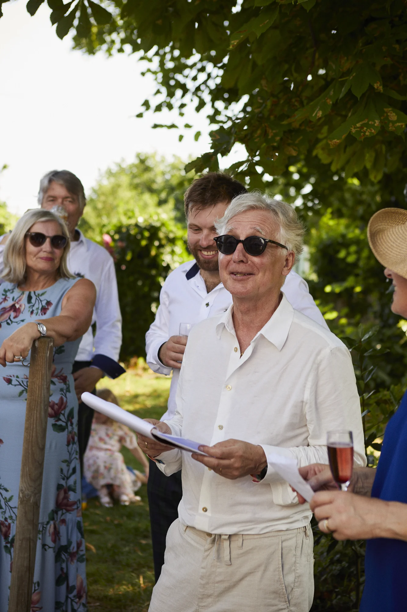 A group of people outdoors, standing under a tree, applauding and smiling, with one person reading from a notebook. They are dressed in summer clothes, some wearing sunglasses, with glasses of a pink beverage in hand, at an outdoor gathering or celeb