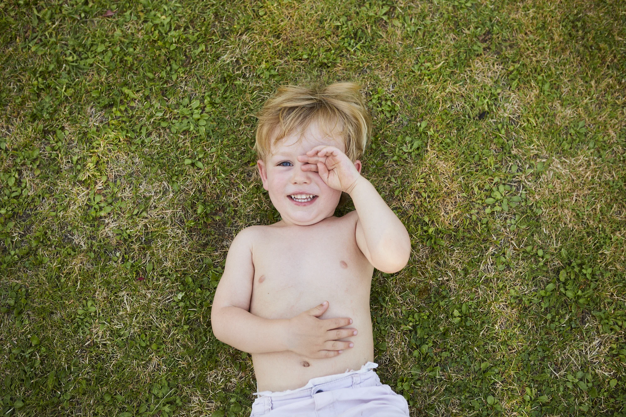 A shirtless young boy with light brown hair and blue eyes lying on grass, smiling, with one hand covering his eye.