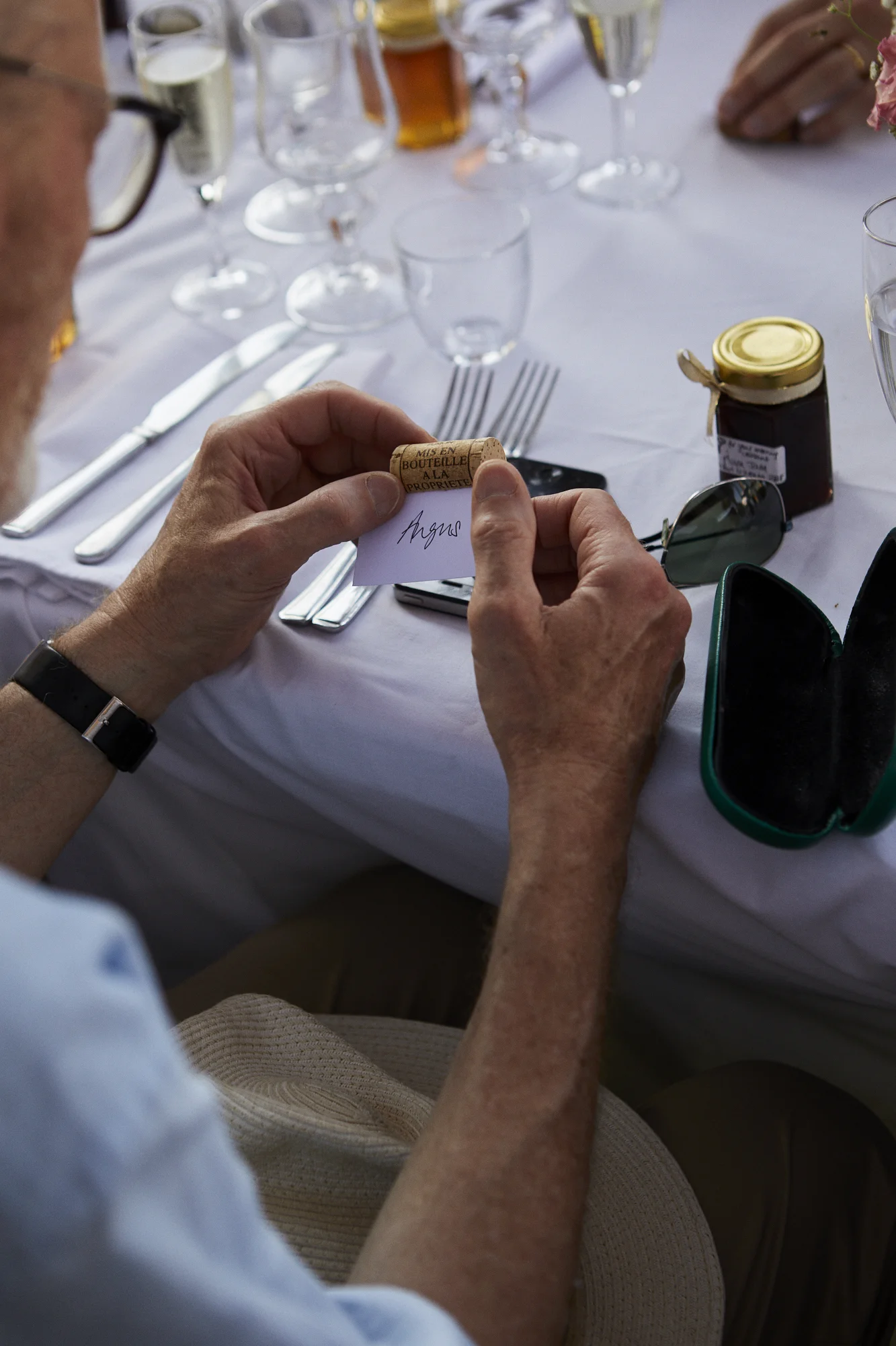 A person holding a wine cork and a small piece of paper at a formal dining table set with glassware, silverware, and a jar of jam.