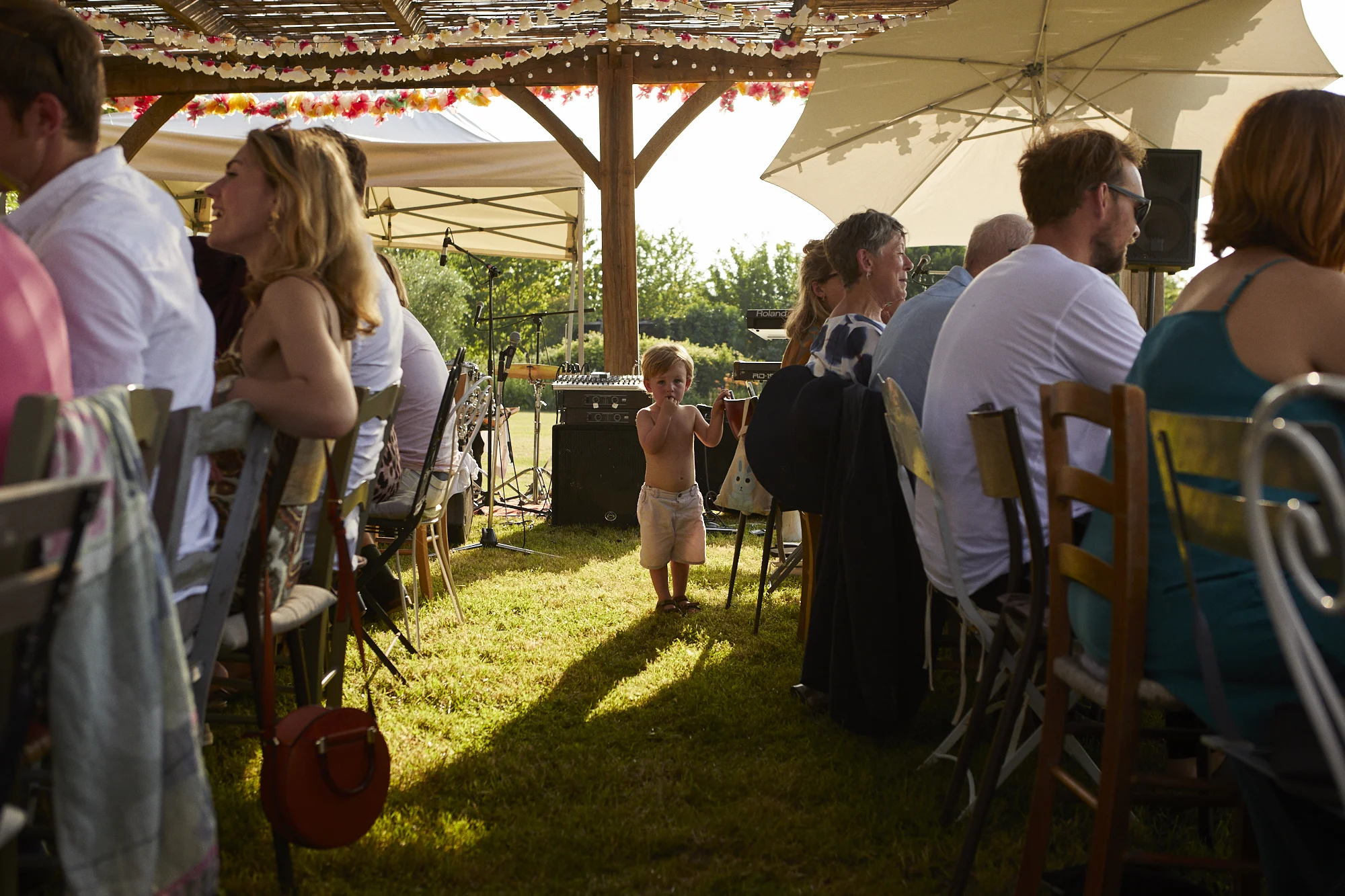 A young boy stands shirtless between two rows of seated adults at an outdoor gathering, with a decorated canopy overhead and musical instruments in the background.