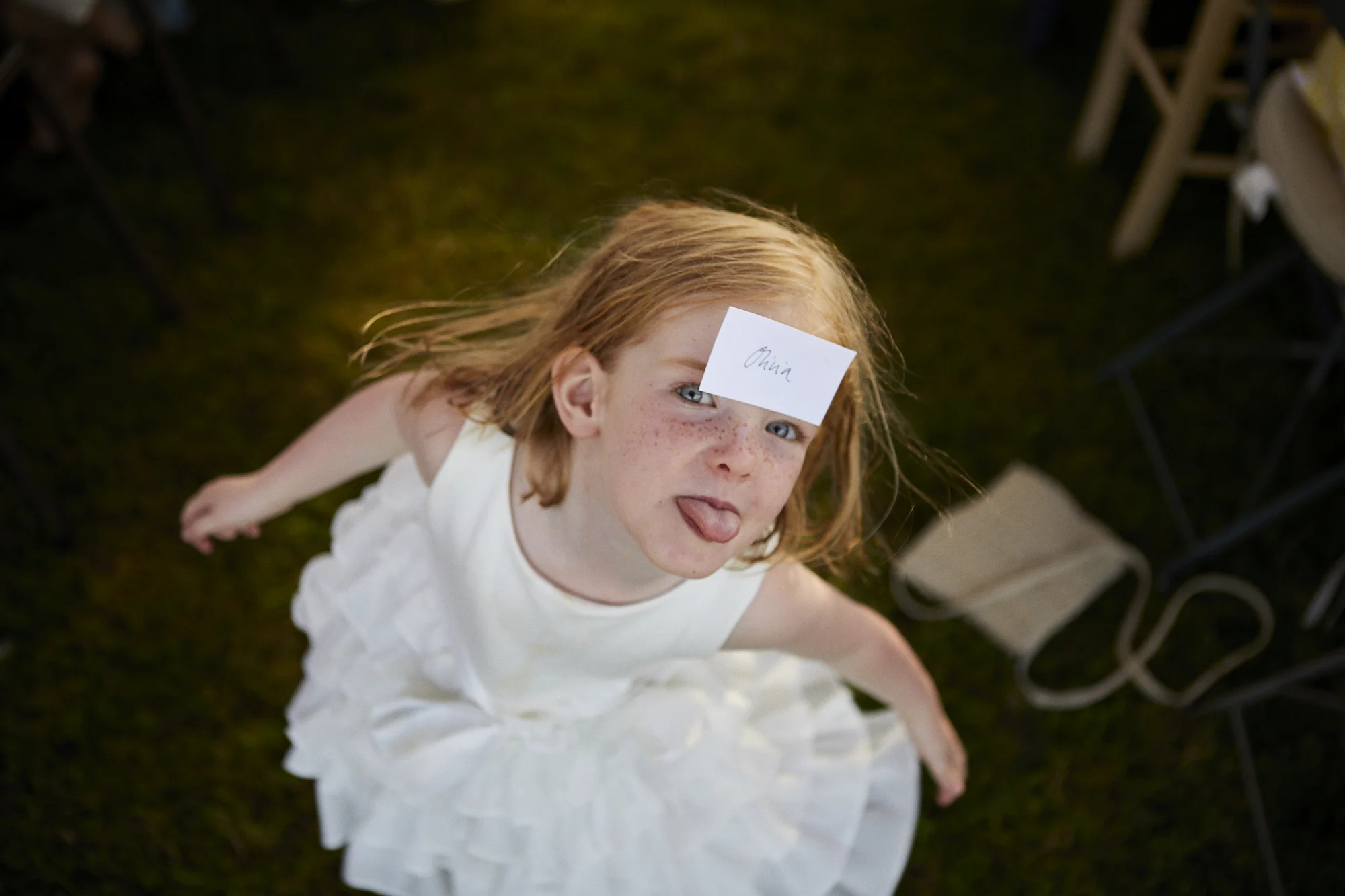 A young girl with red hair and freckles sticking out her tongue, wearing a white dress, with a note on her forehead that says "China."