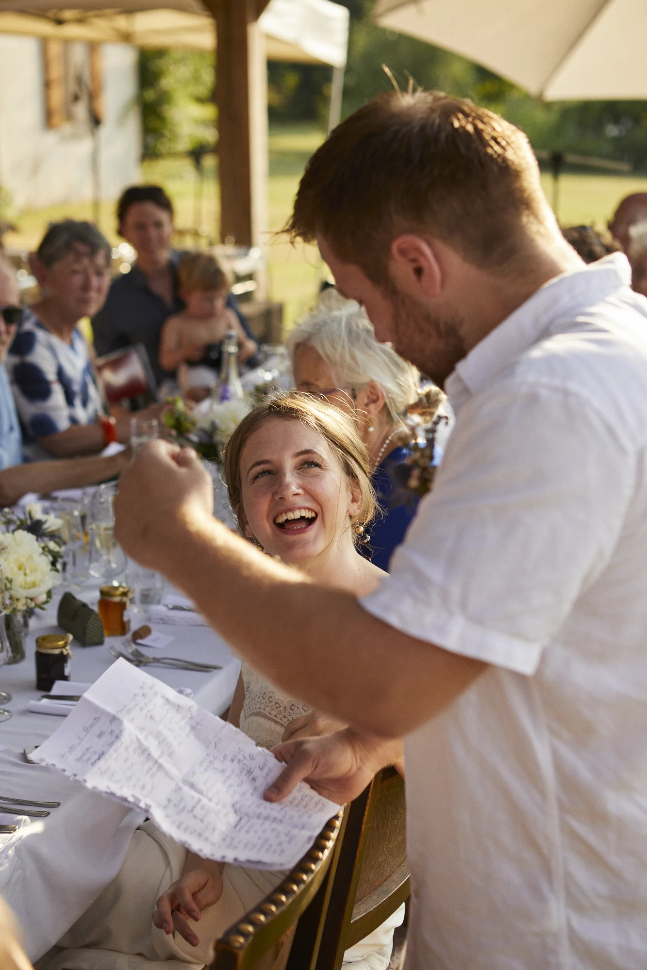 A joyful young woman with red hair and a white dress laughing at an outdoor gathering, seated at a table with other guests, while a man in a white shirt shows her something on a paper.
