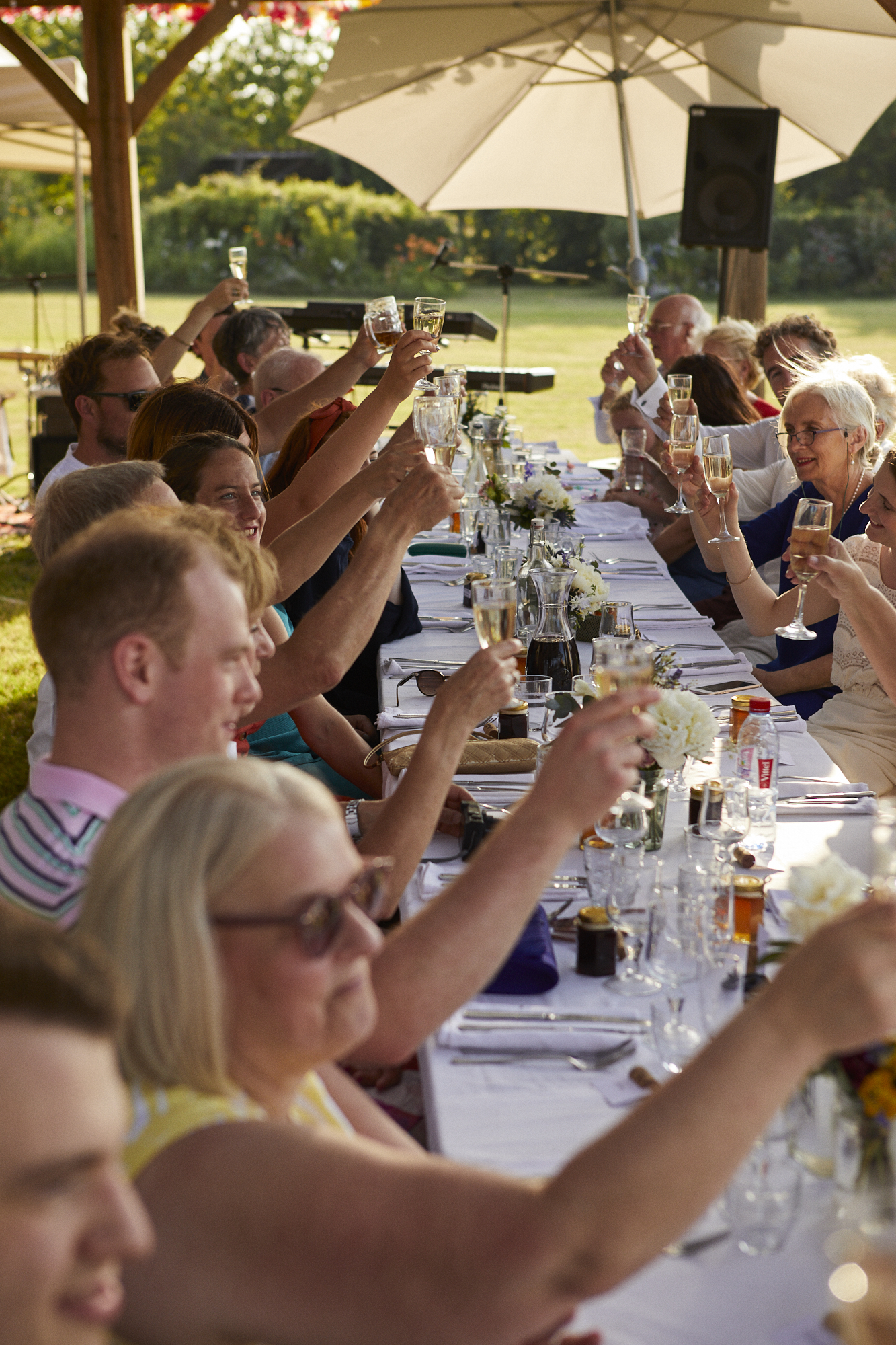 People gathered at an outdoor celebration dining table, raising glasses in a toast under a large umbrella.