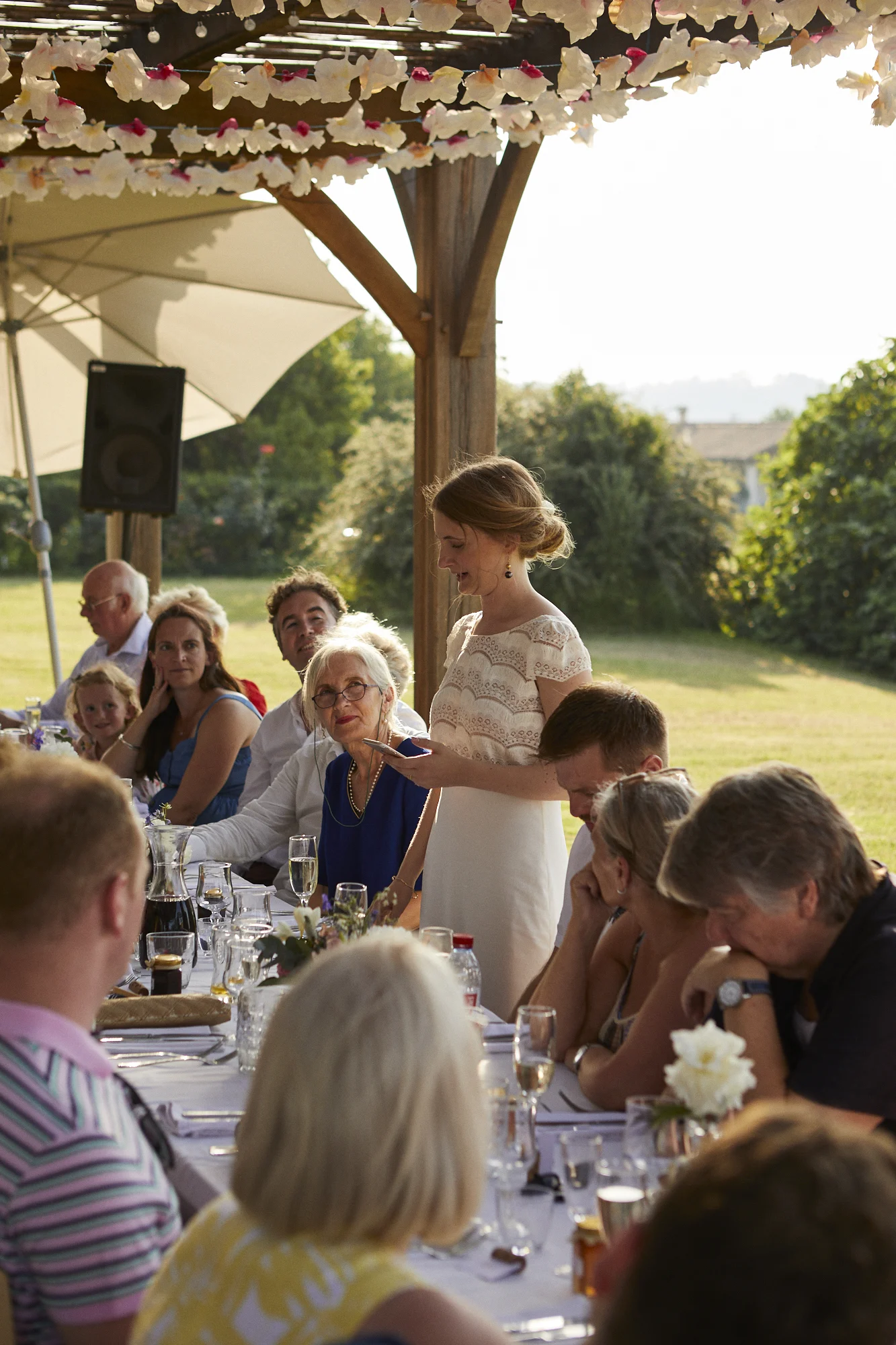 A woman standing at an outdoor event table, speaking or giving a toast, with people seated around her.
