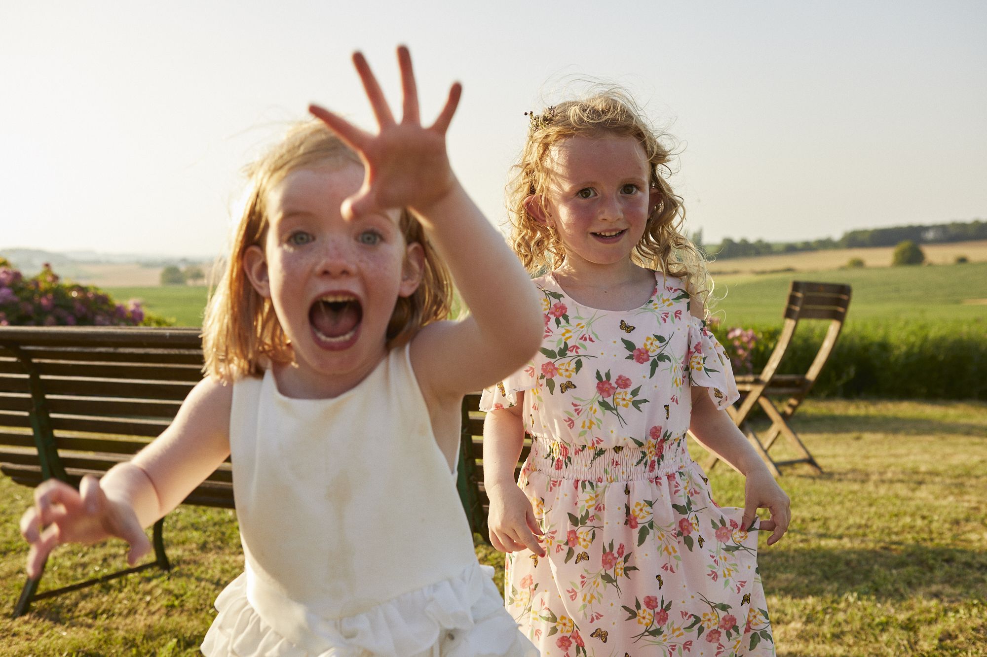 Two young girls with curly red hair playing outdoors on a sunny day, one girl is in the foreground with her mouth open and hand reaching towards the camera, the other girl is in the background smiling, with a wooden bench and green fields behind them