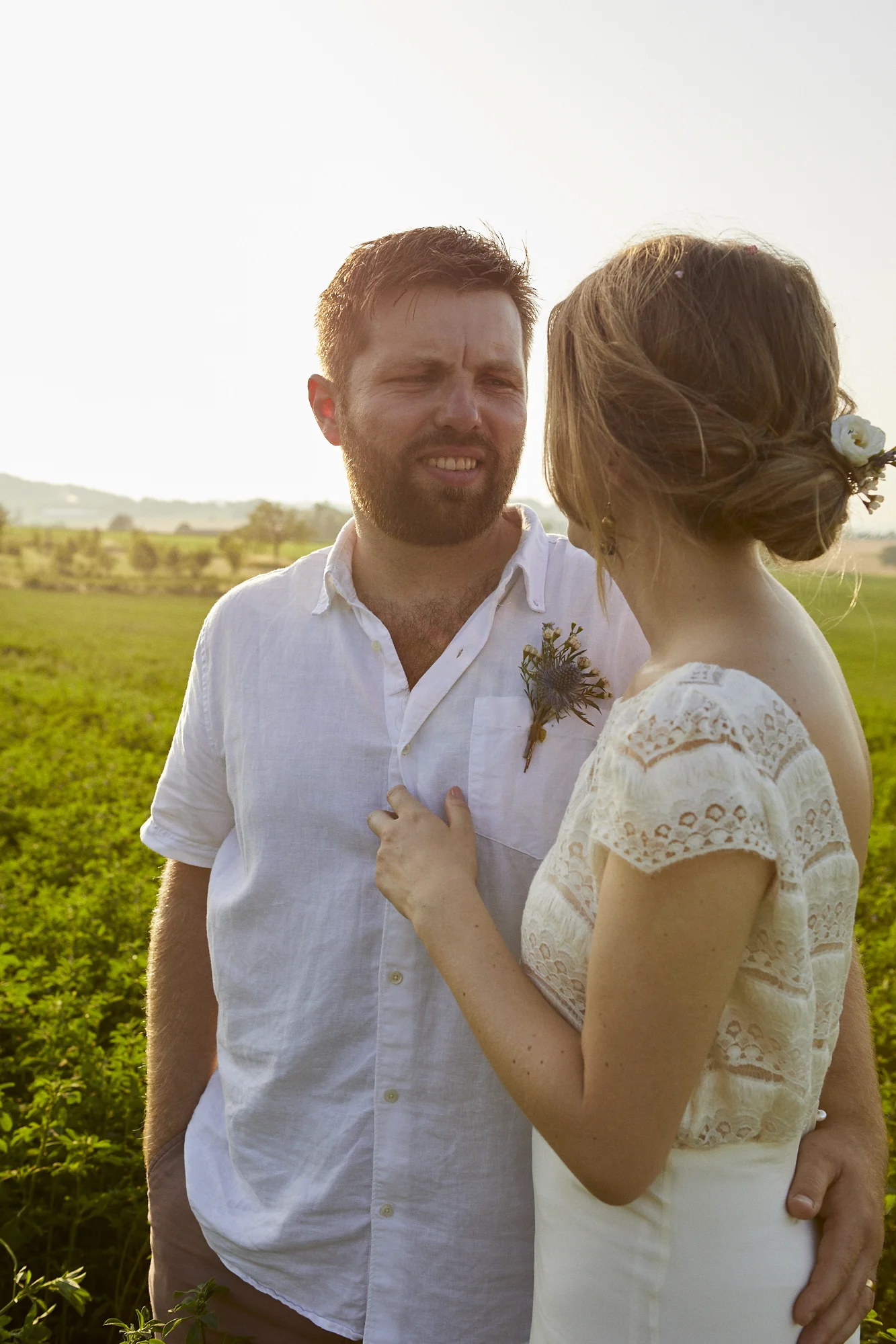 A man and woman in a field during sunset, dressed in white, close together, with the man showing a slightly puzzled or emotional expression.