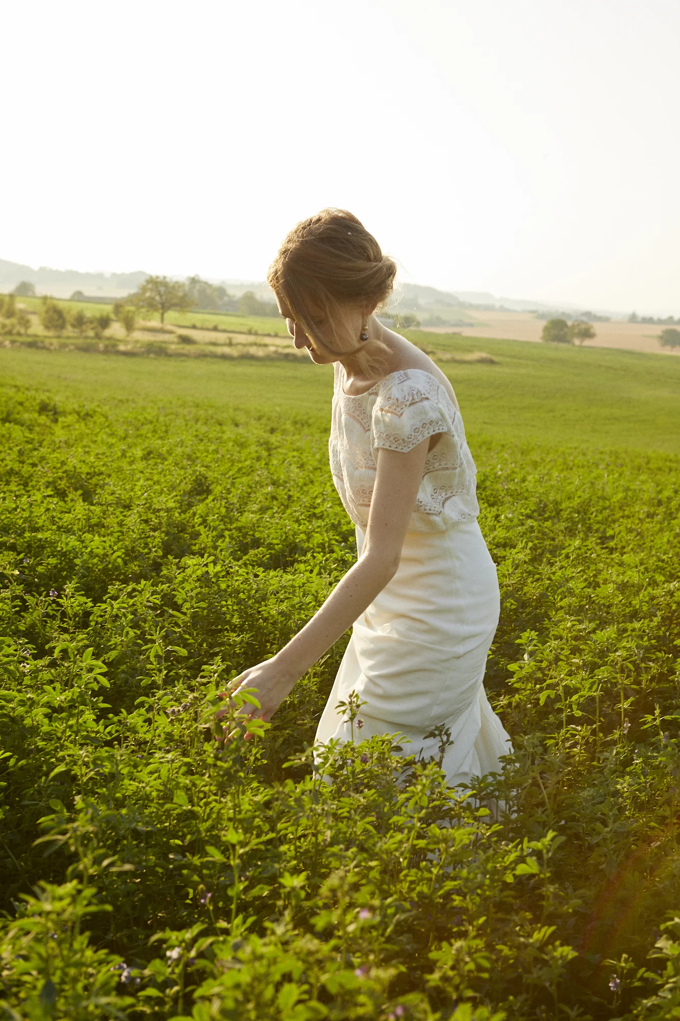 A woman in a white lace top and long white skirt standing in a green field, gently touching the plants, with a scenic countryside background during sunlight.