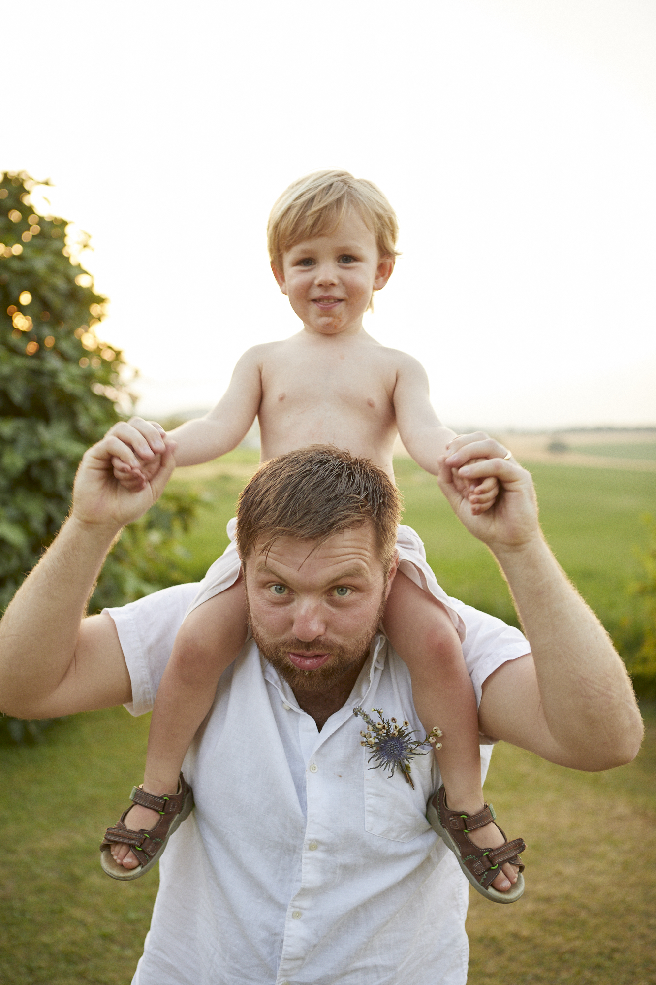 A man with a beard carrying a young boy on his shoulders outdoors during sunset, with green fields and trees in the background.