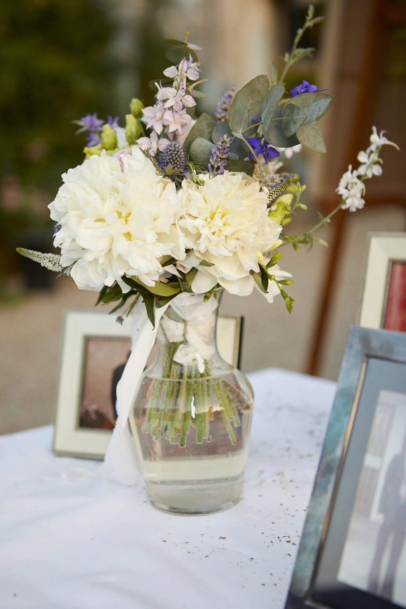 A floral arrangement in a clear glass vase containing white peonies, purple flowers, eucalyptus leaves, and other greenery, placed on a white table with framed photos in the background.