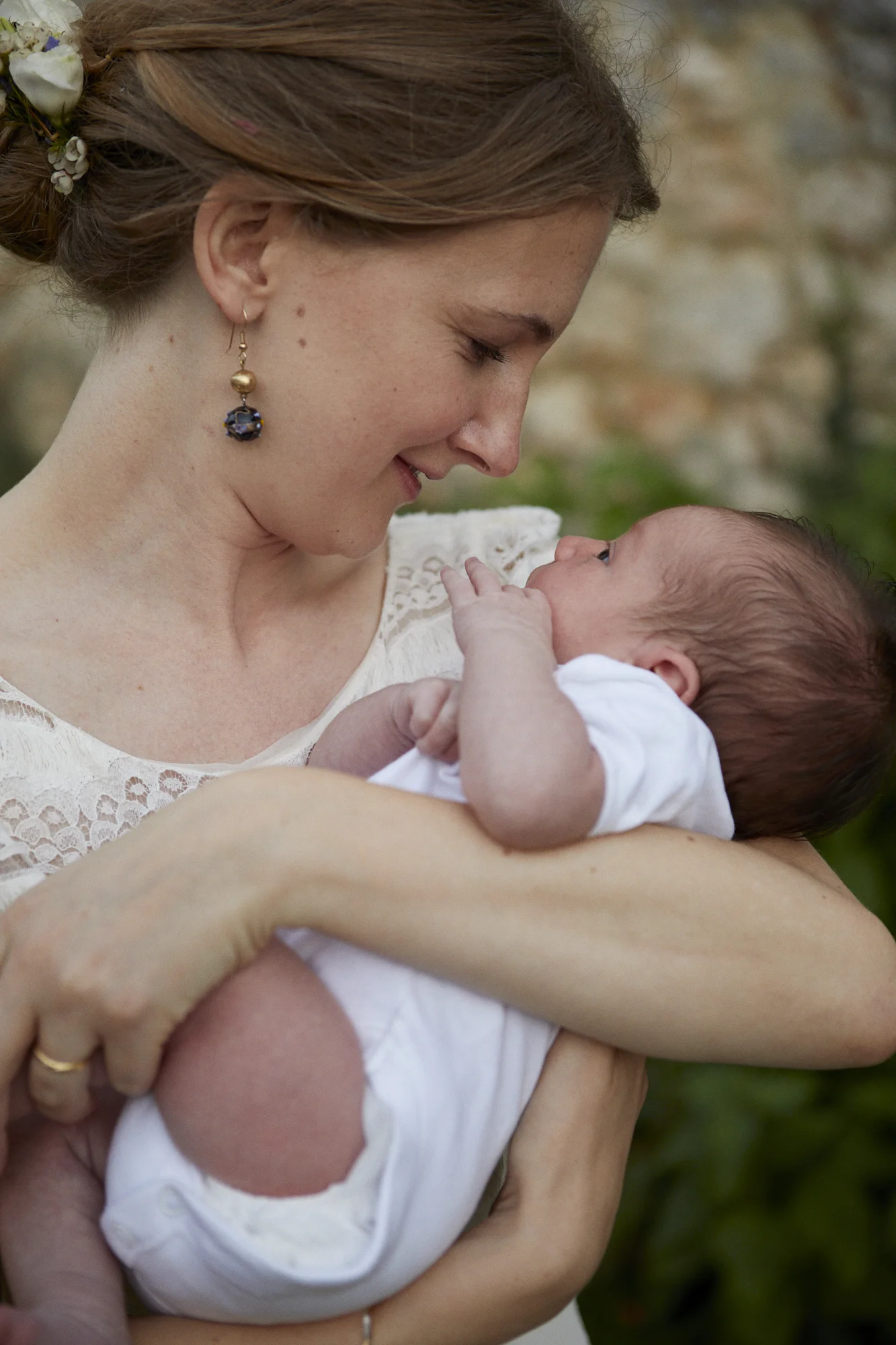 A woman holding a newborn baby close to her face, smiling with eyes closed, outdoors with a blurred stone wall background.