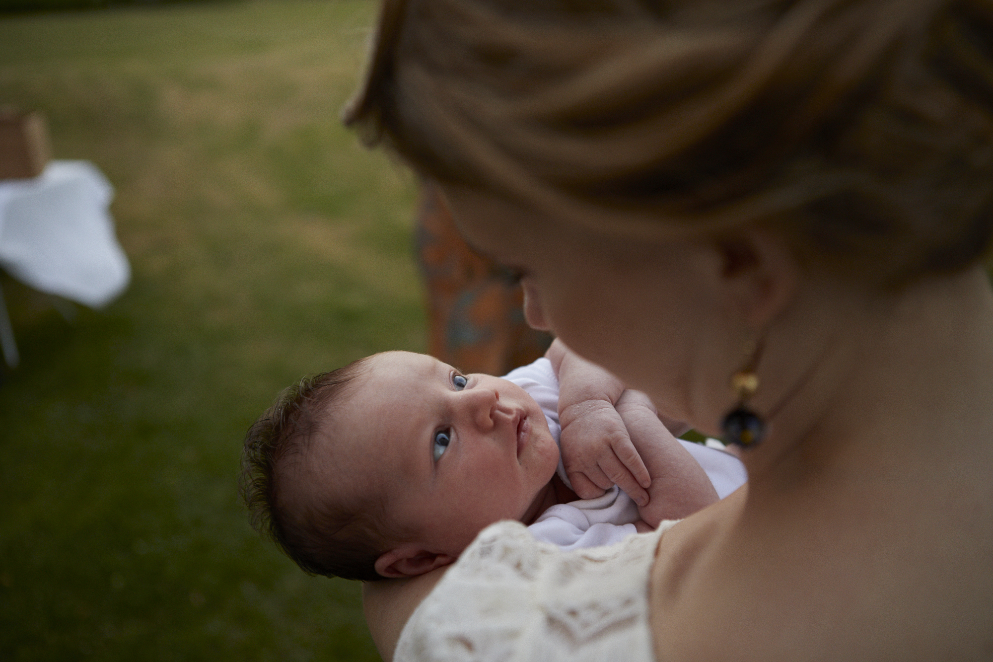 A woman with light brown hair gazes affectionately at a newborn baby with blue eyes as she holds the baby close to her. The scene is outdoors on grass with some furniture in the background.