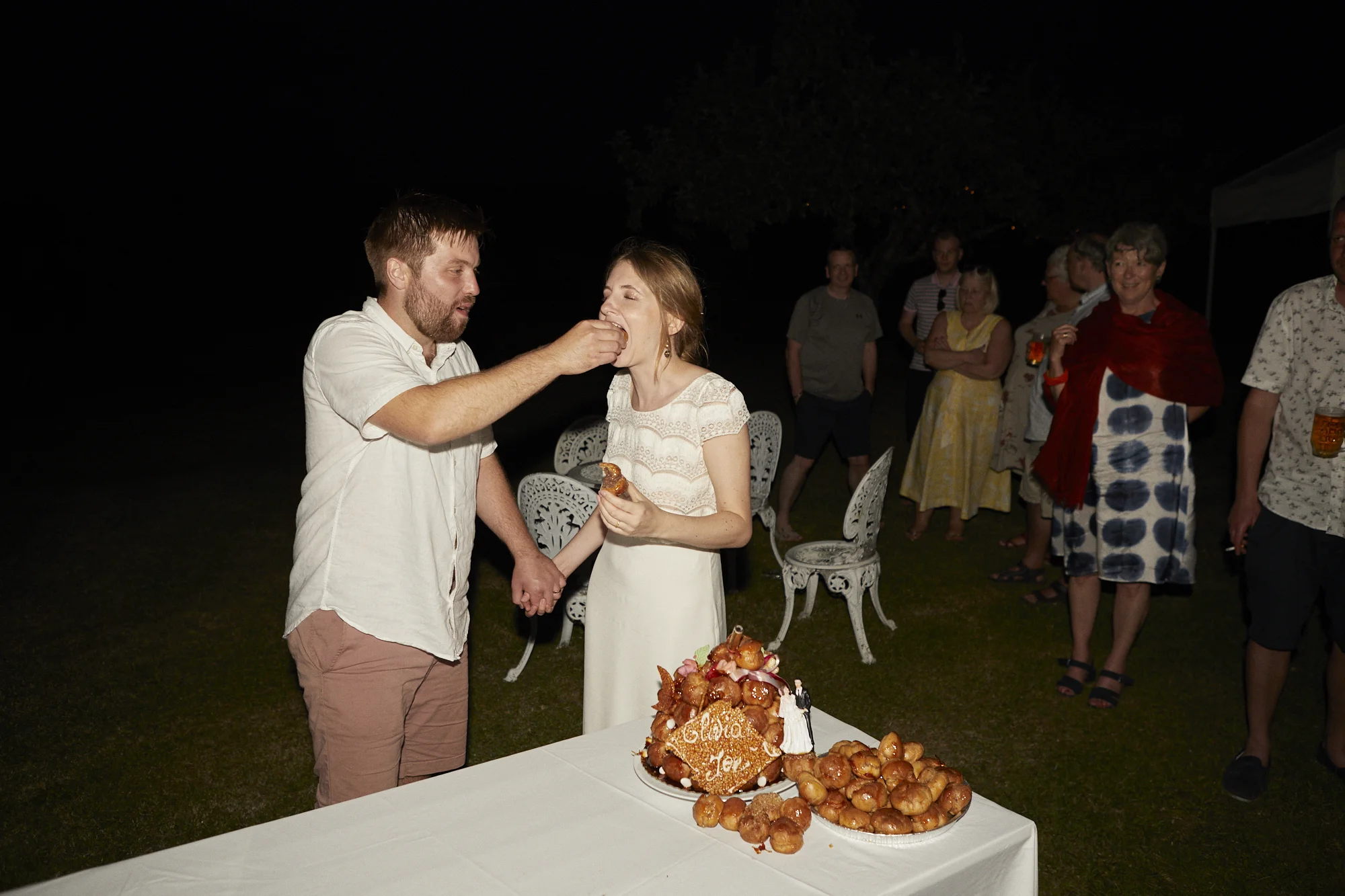 A couple is celebrating their wedding outdoors at night, with the groom feeding cake to the bride while holding hands. A cake decorated with cream and strawberries is on the table in front, with several cream puffs surrounding it. Family and friends 