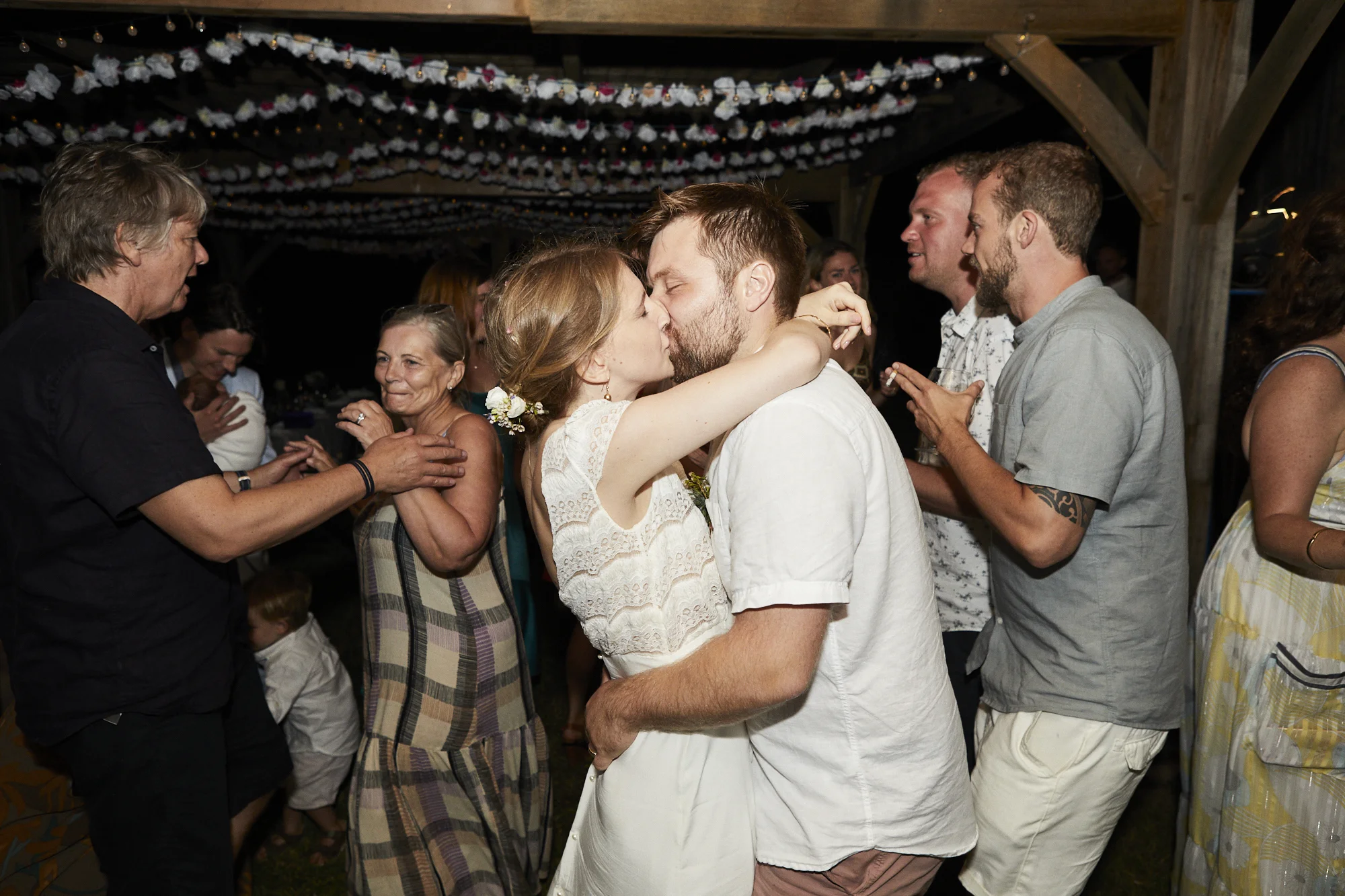 A couple sharing a kiss at a wedding reception surrounded by family and friends.