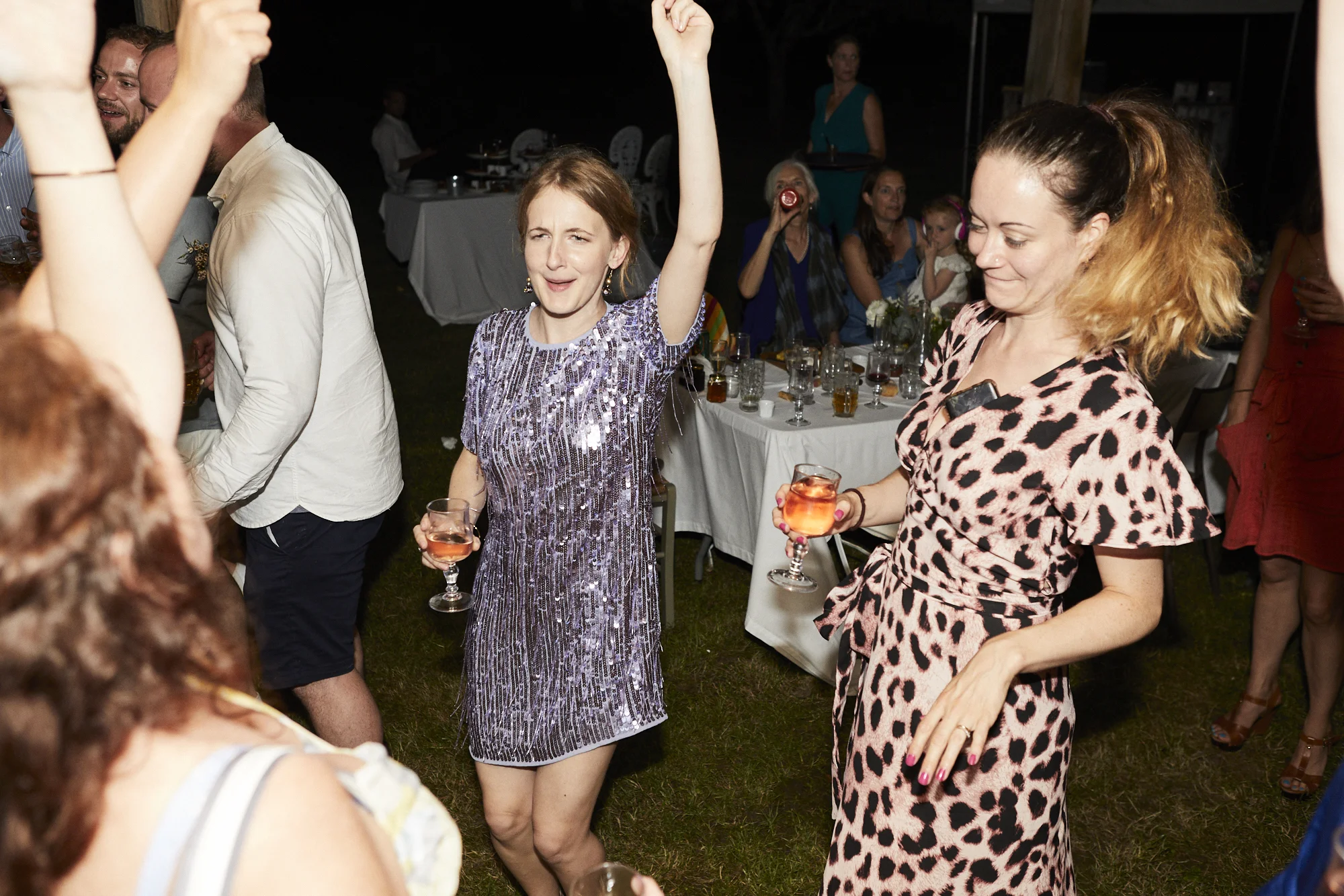 People dancing and enjoying drinks at a social gathering or party, with a woman wearing a purple, sequin dress and another woman wearing a leopard print dress holding glasses of pink beverage.