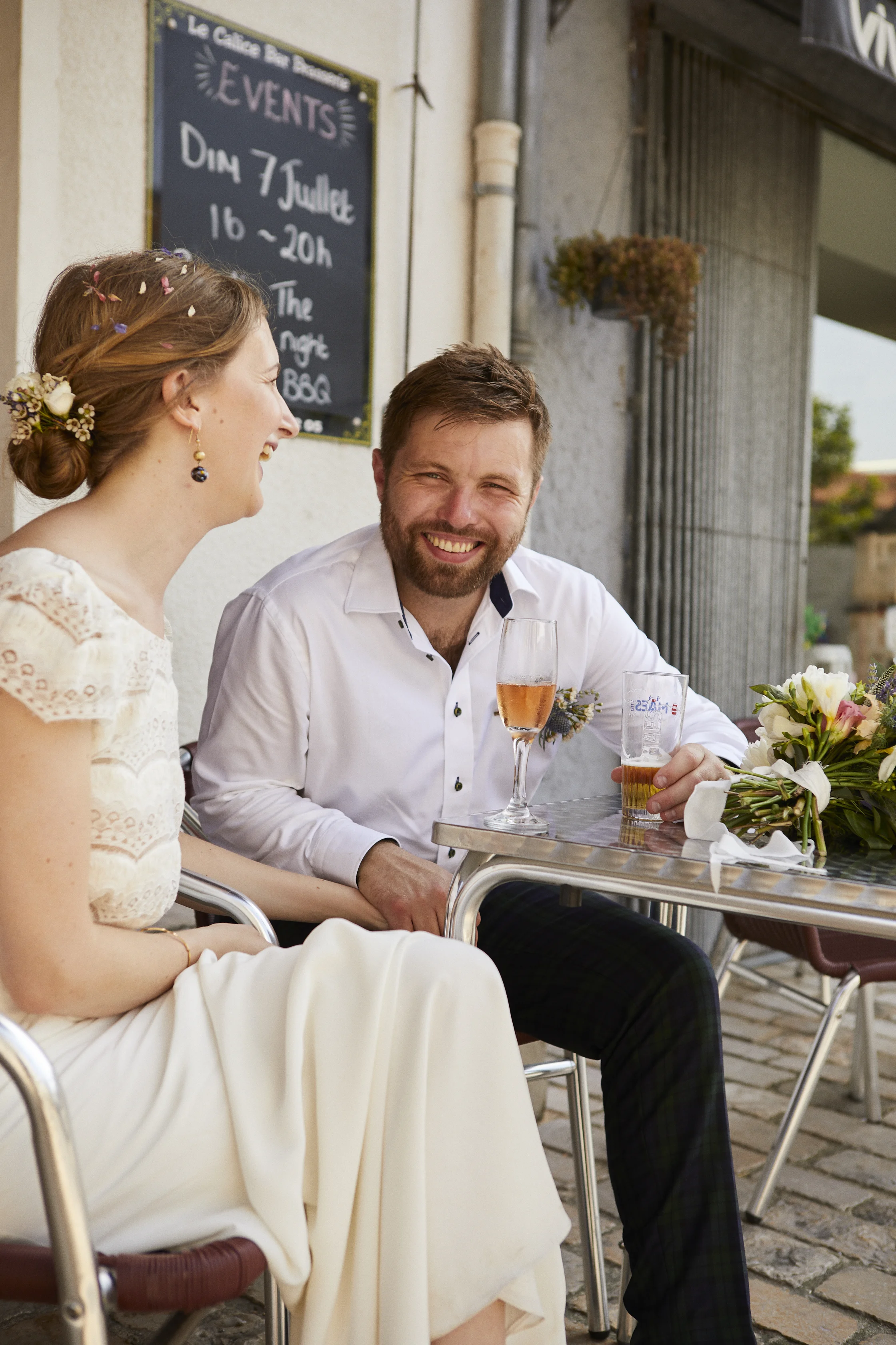 A man and woman sitting at an outdoor cafe, enjoying drinks and engaging in conversation with smiles, with a chalkboard sign behind them and flowers on the table.